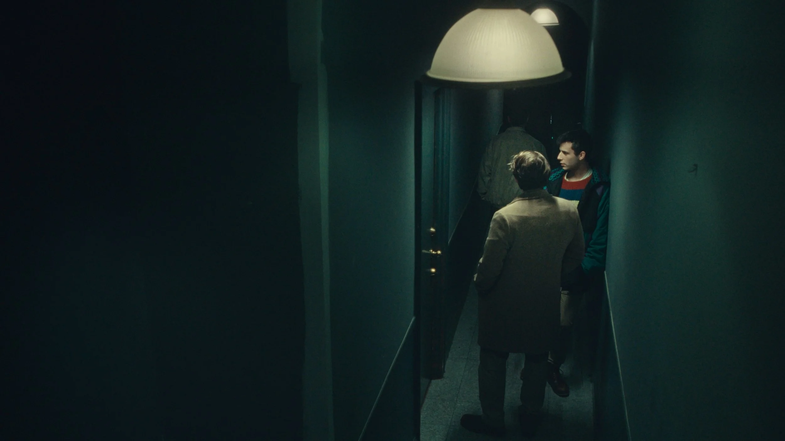 Three people standing and talking in a dimly lit hallway near a big ceiling lamp, with one person facing away from the camera.