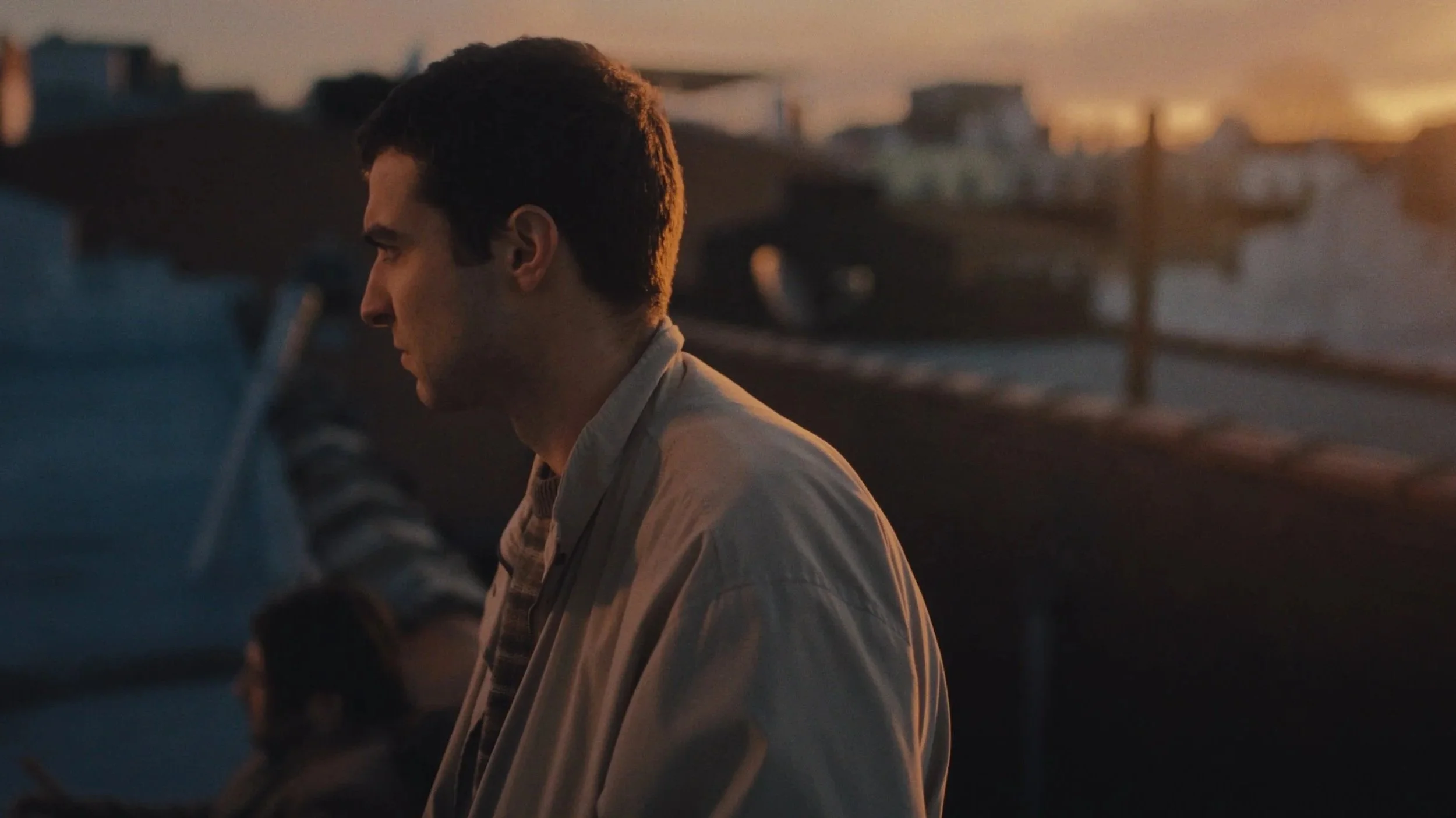 A man in profile sits outdoors during sunset, on a roof with  buildings in the background.