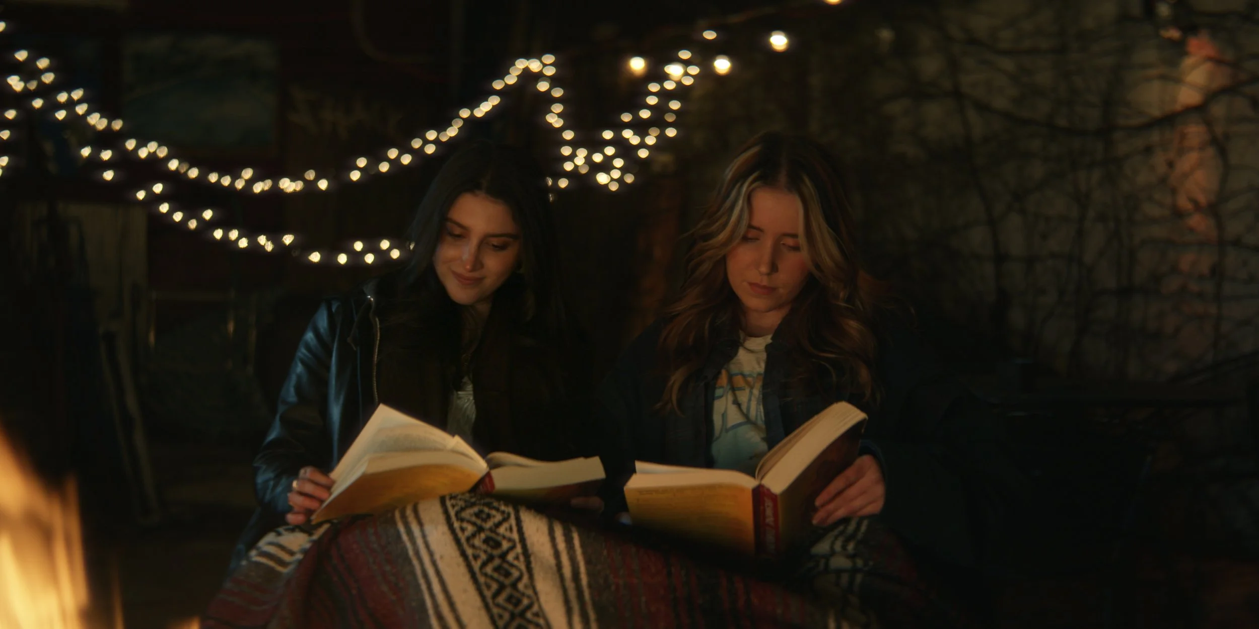 Two women sitting together at night outdoors, reading books, with string lights in the background.