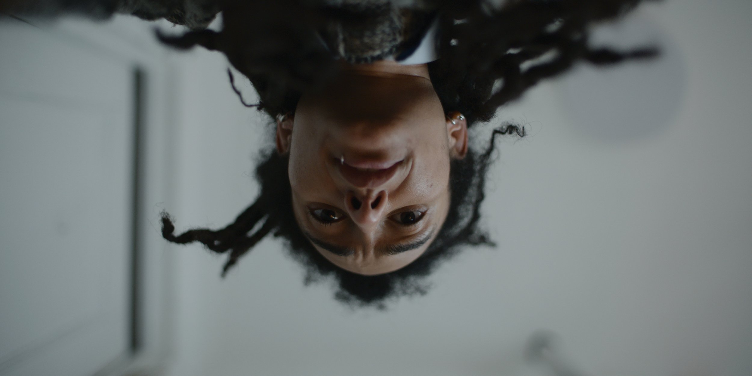 Upside-down shot of a woman with dark curly hair, wearing earrings, looking at the camera with a slight smile.