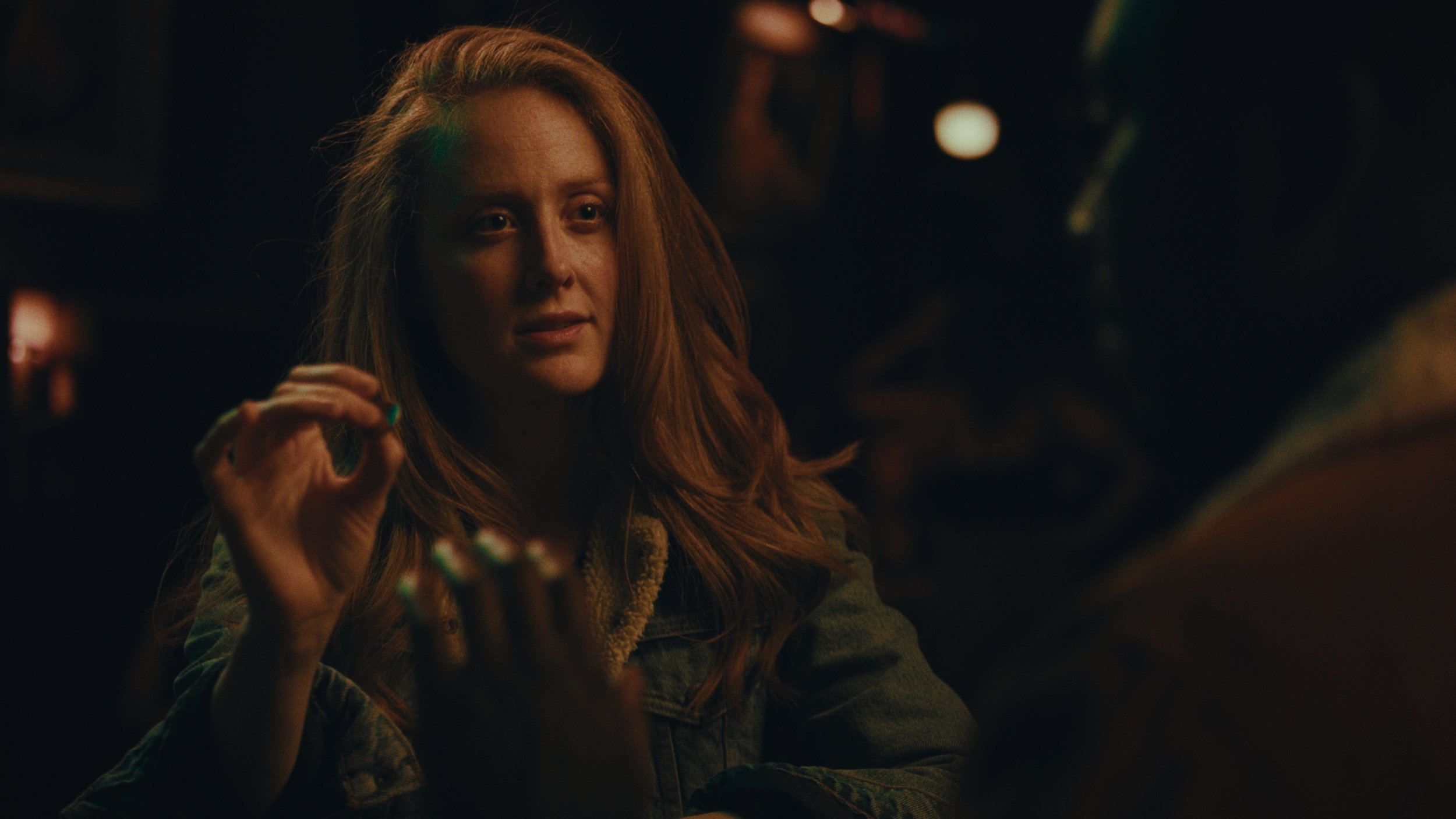 A woman with long red hair, wearing a denim jacket, talking in a dimly lit environment.