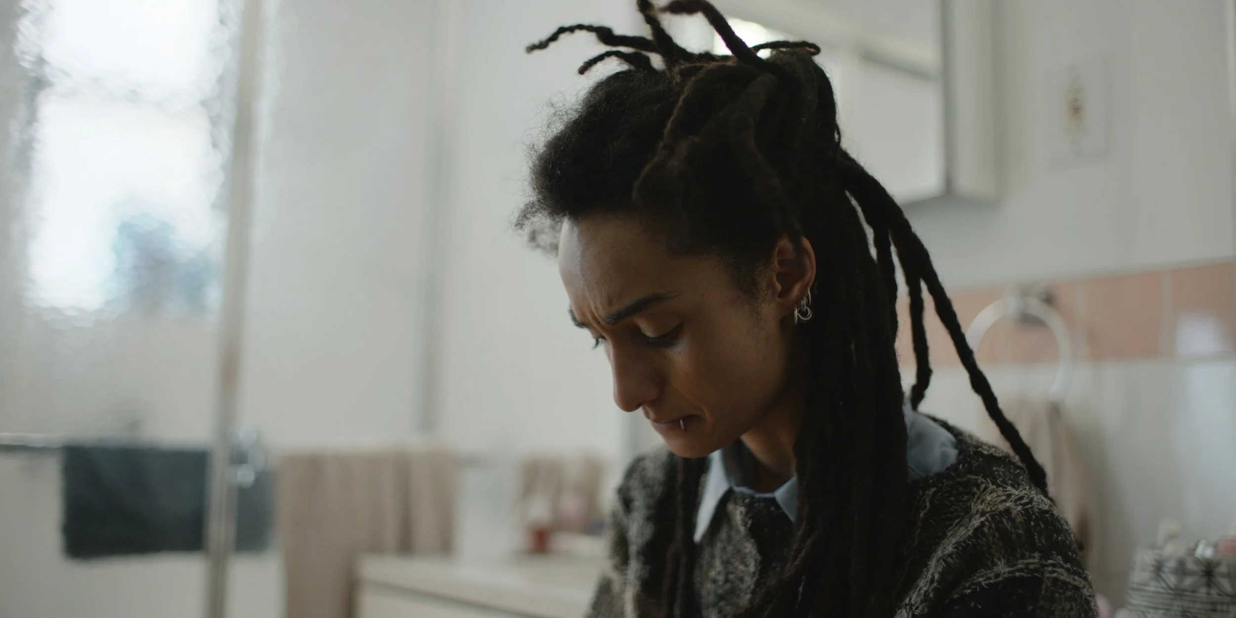 A young woman with dreadlocks and earrings looking down thoughtfully in a room with a white wall and window.