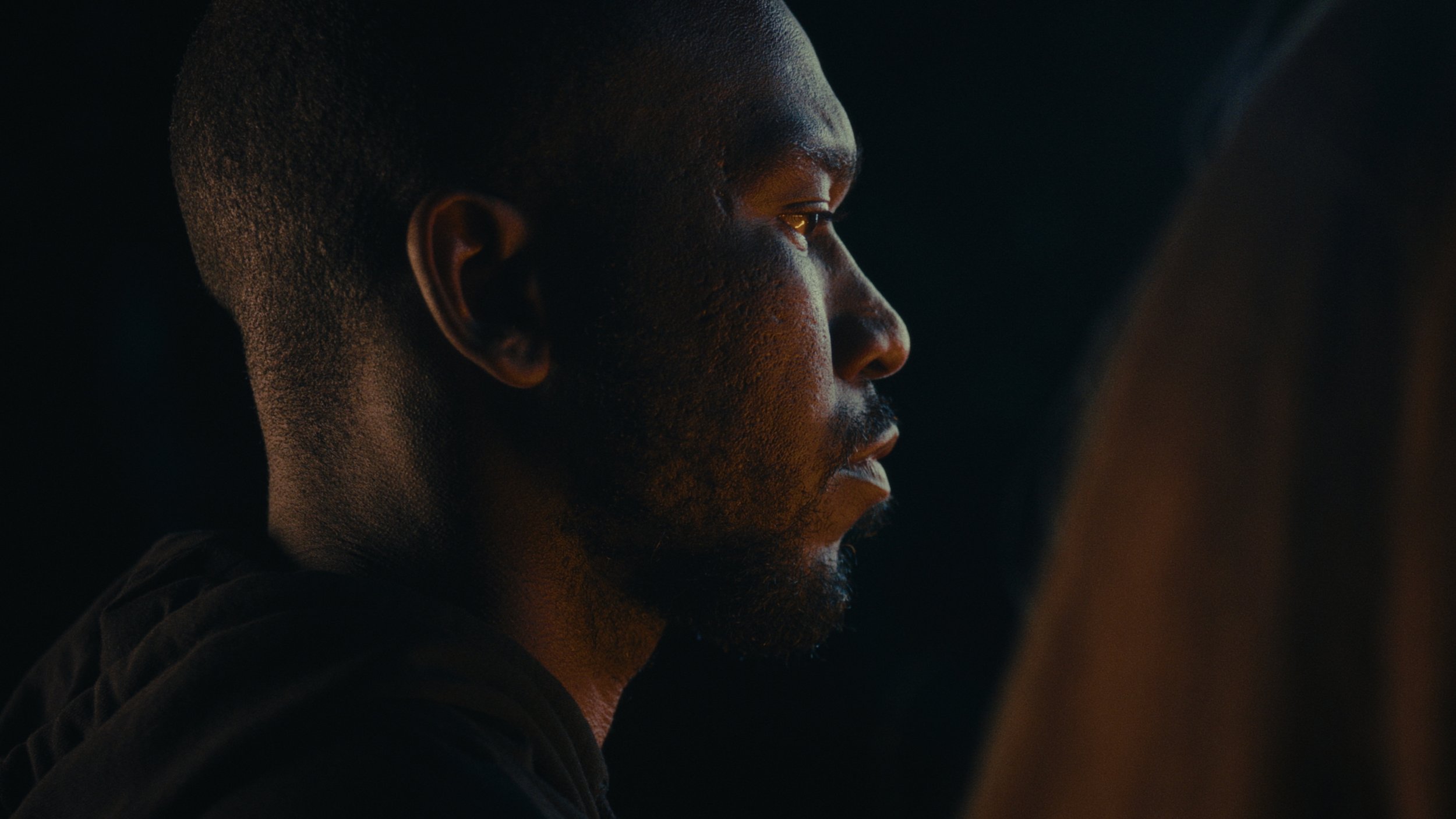 A side profile of a man with short hair and a beard, looking intently forward in a dark setting with subtle lighting highlighting his facial features.