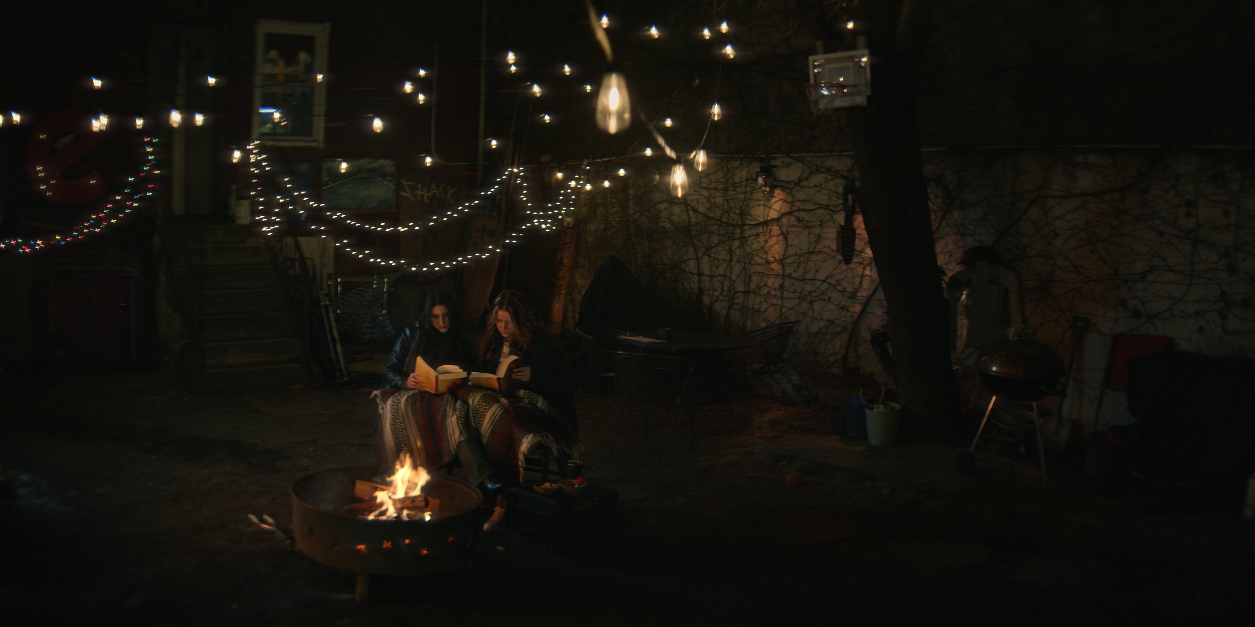 Two women sitting around a firepit in a dark backyard, reading books with string lights hanging overhead.