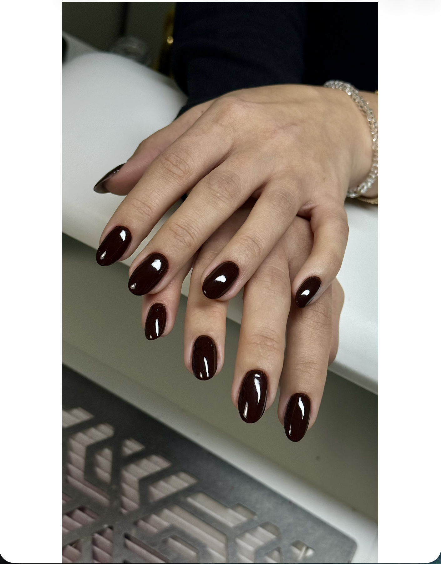 Hands with manicured nails painted in dark burgundy, wearing a bracelet and a dark long-sleeve shirt, resting on a white cylindrical surface.