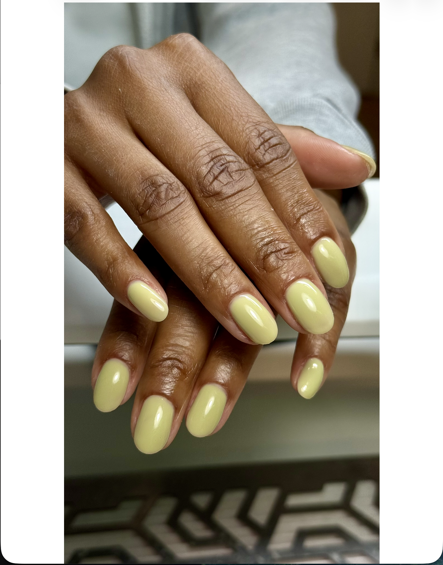 Close-up of a person's hands with manicured nails painted in light yellow, resting on a surface. The person has a medium brown skin tone and visible wrinkles on the hands.