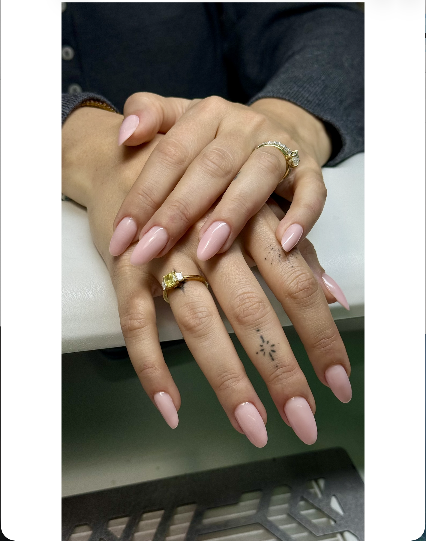 Close-up of two hands with manicured nails and rings, resting on a white surface. One hand has a tattoo on the finger. The background includes a person wearing a dark jacket.