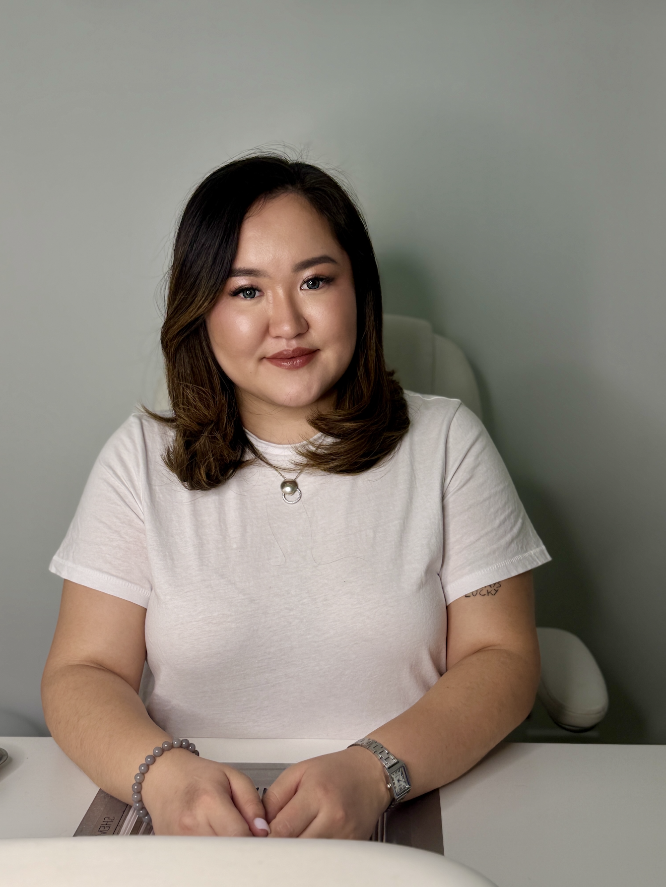 A woman with shoulder-length brown hair, wearing a white shirt, sitting at a desk with hands clasped. She has a silver watch, a bracelet, and a necklace with a pearl, and is smiling gently in front of a pale green wall.