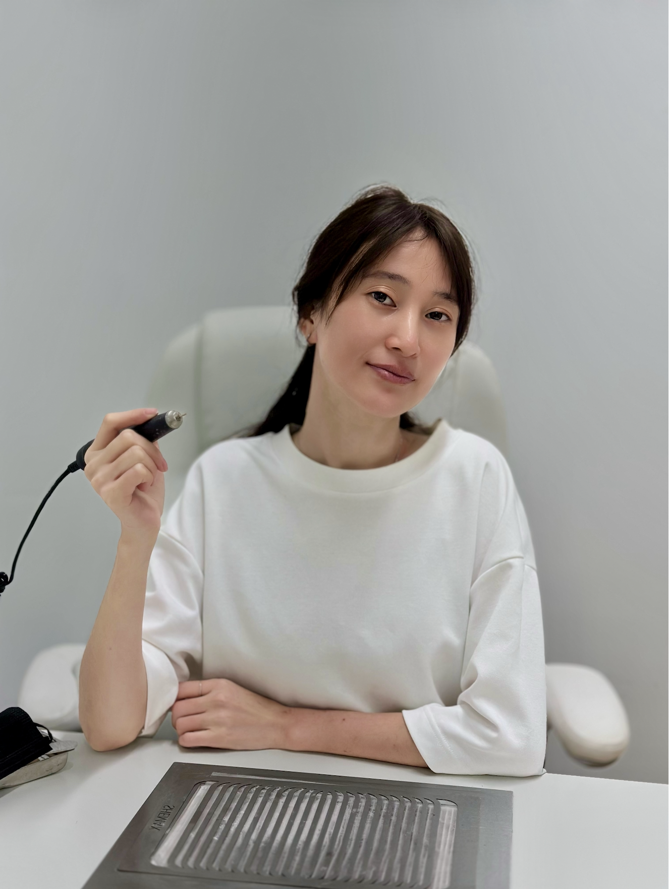 A woman in a white shirt sitting at a table, holding a nail drill, with a gray wall in the background.