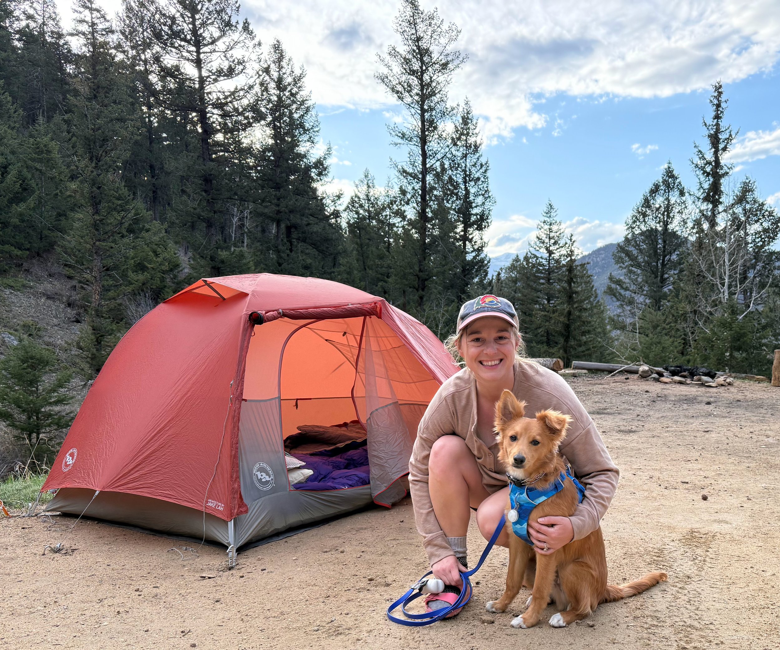 Rachel McCarl is wearing a Colorado hat and crouching on the ground, holding her reddish border collie mix, Otto, who is wearing a blue harness. There is a red tent with the tent door askew. In the background are pine trees and scenic mountains.