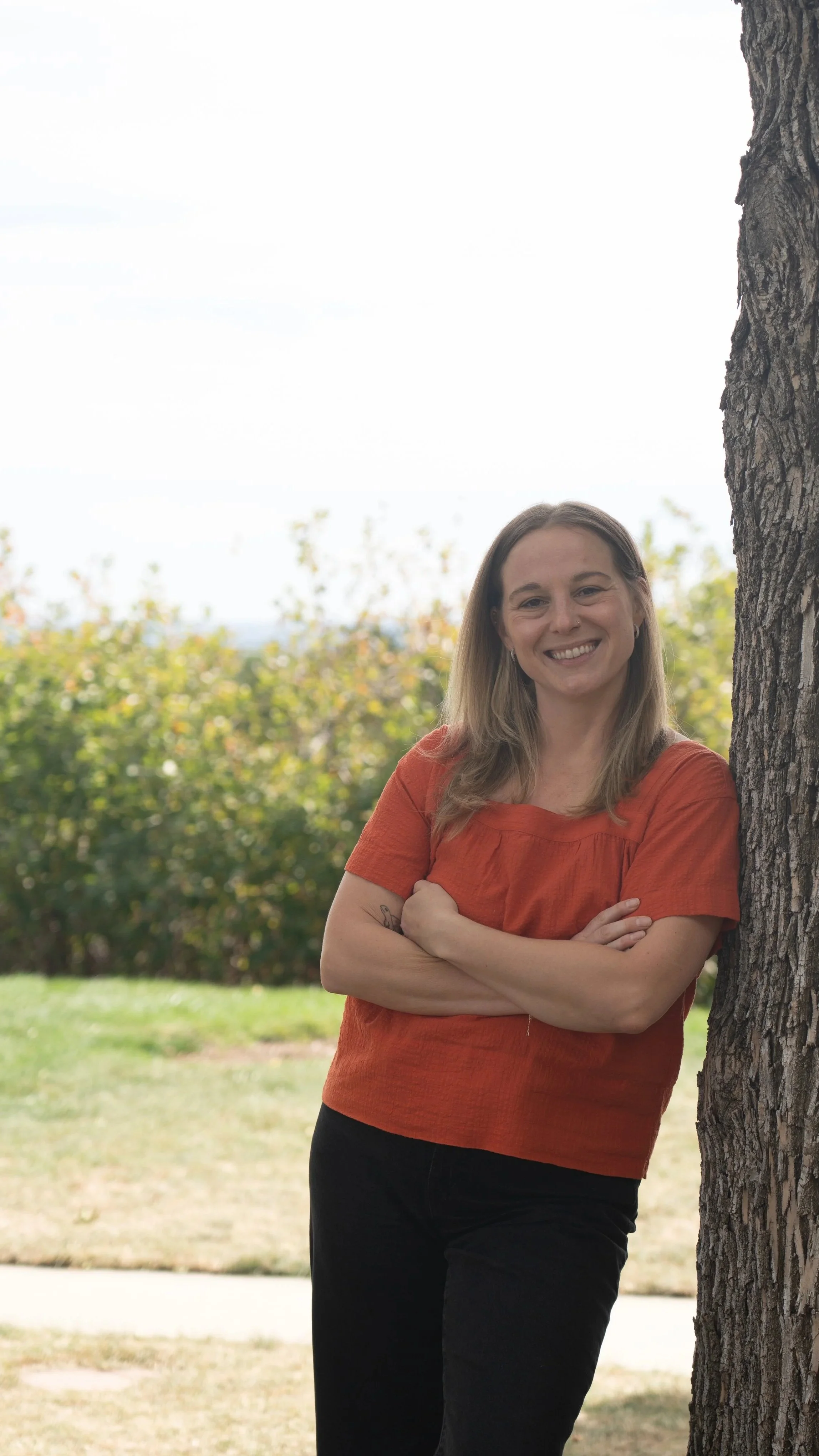 A woman with light brown hair, wearing an orange top and black pants, standing outdoors next to a tree with arms crossed, smiling at the camera.