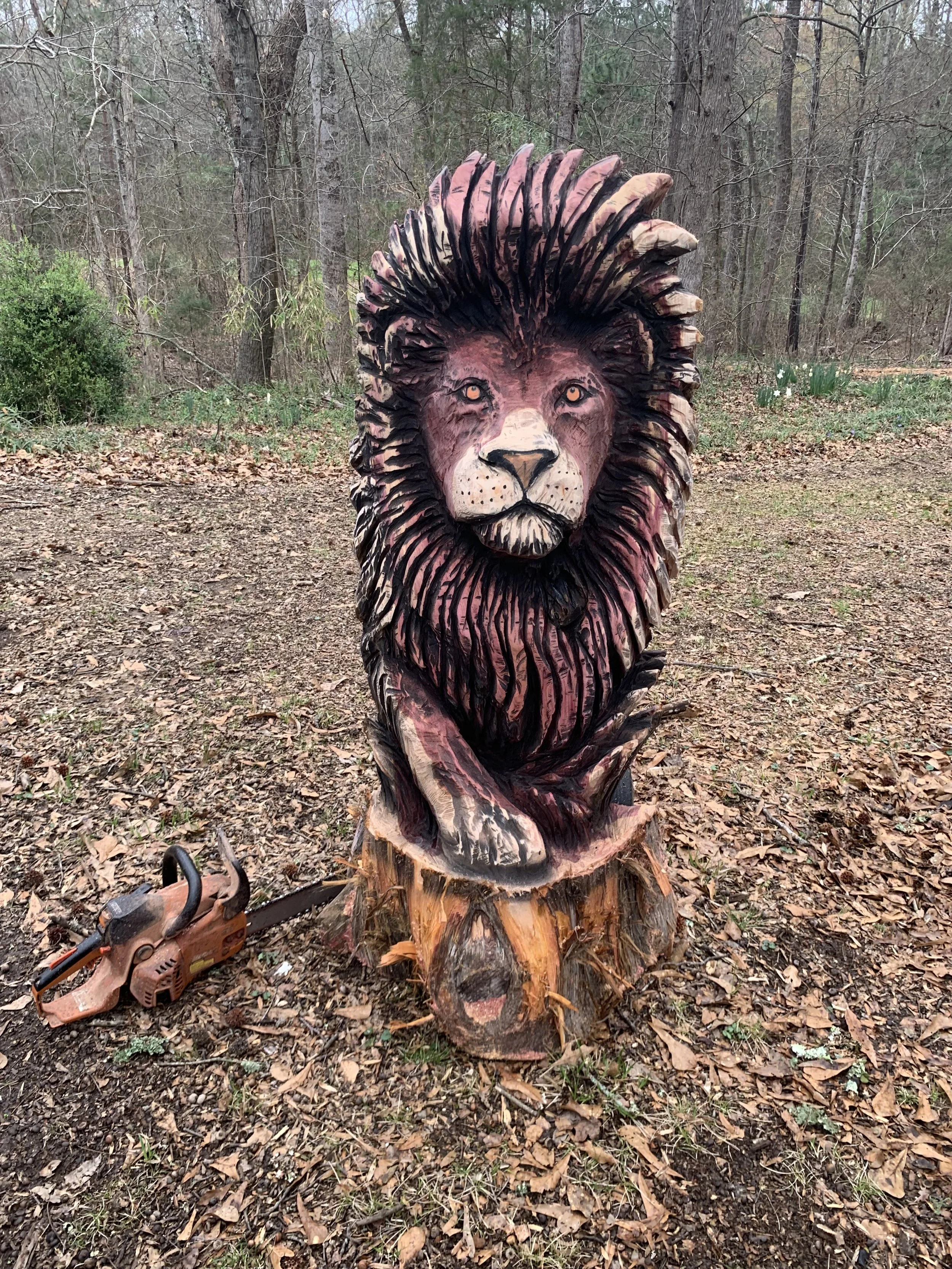 A hand-carved wooden sculpture of a lion's head with a detailed mane, mounted on a tree stump in an outdoor forest setting with leaves and trees in the background.