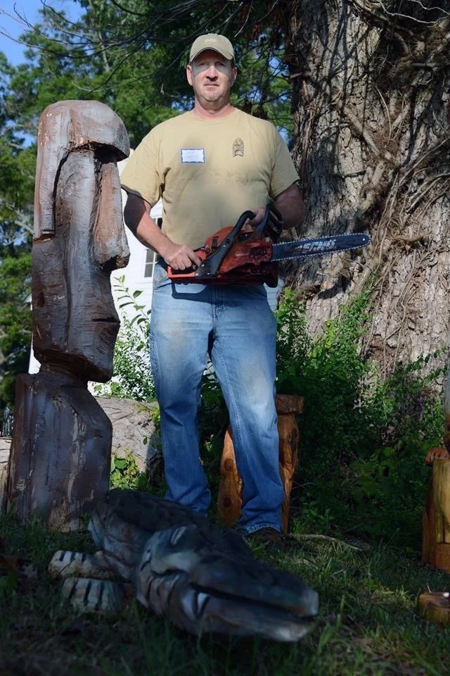 Man standing outdoors with chainsaw, surrounded by carved wooden sculptures, including a crocodile in the grass, trees in the background.