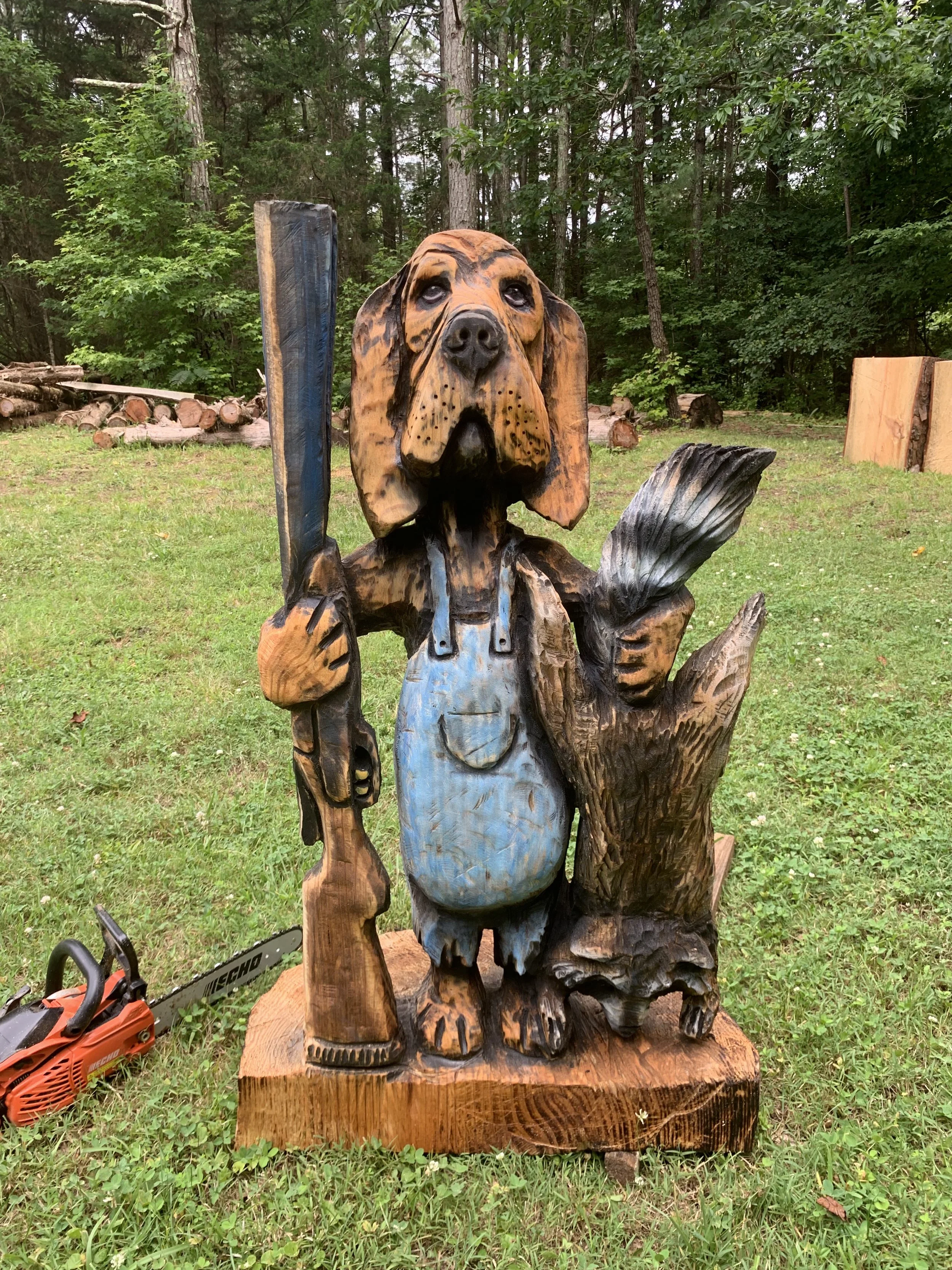Wooden sculpture of a dog holding a chainsaw, surrounded by trees and logs in a forested area.