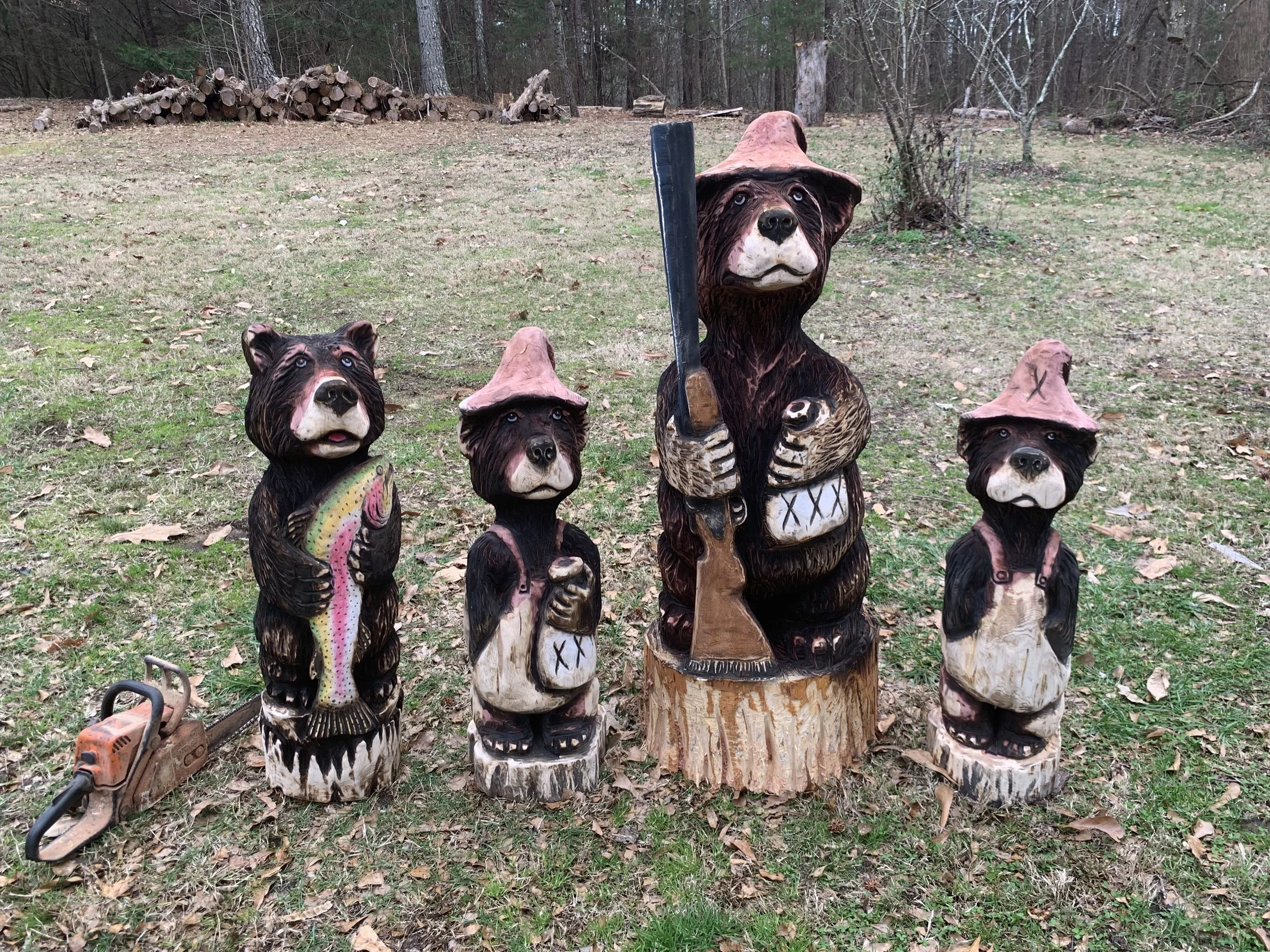 A group of four carved bear statues, each wearing a pink hat, are displayed outdoors in a wooded area on grass with scattered leaves. A chainsaw is visible on the ground to the left of the statues, and in the background, logs are stacked along the tree line.