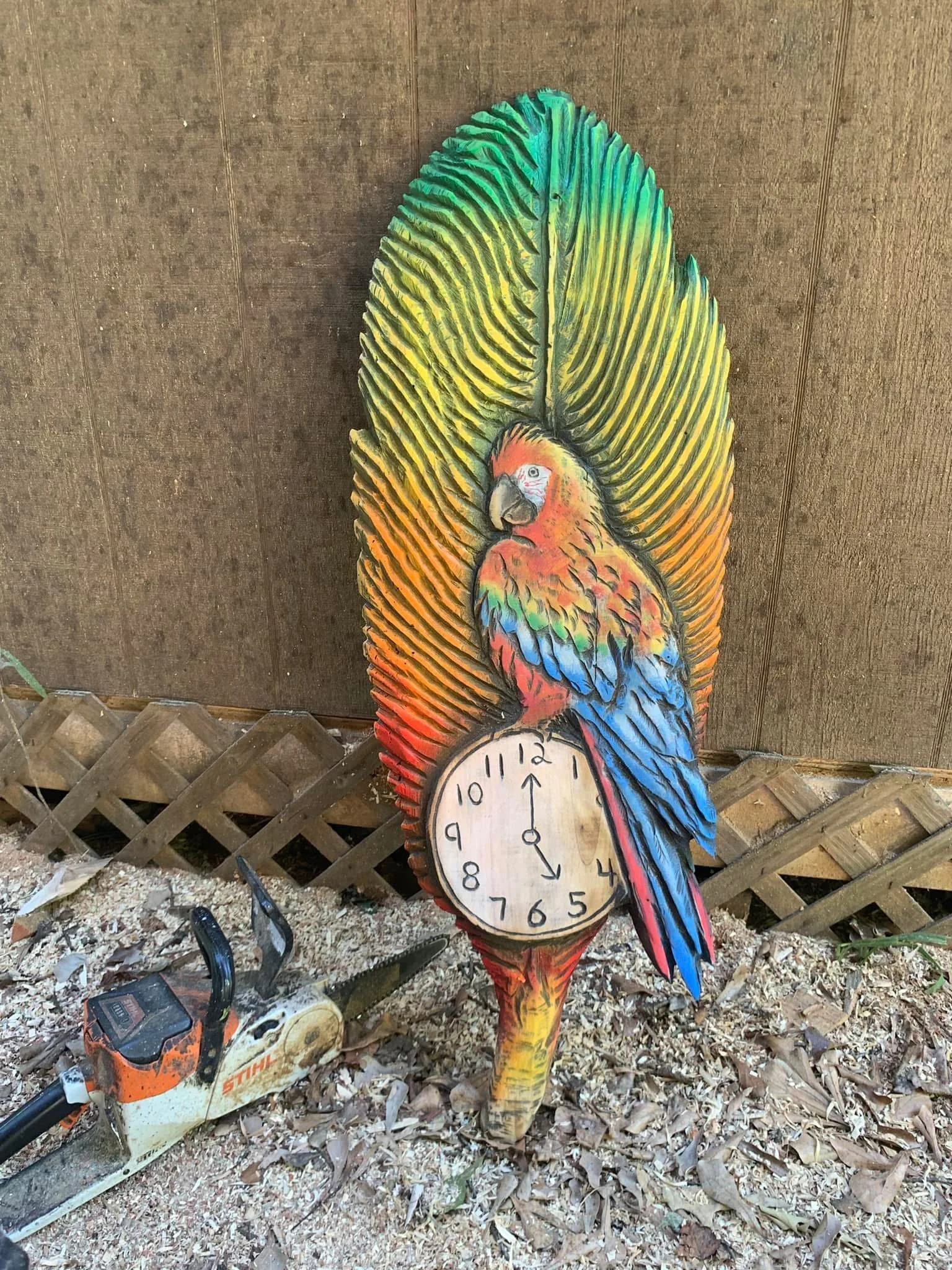 A colorful wooden cutout of a bird with large feathers serves as a clock, set against a wooden wall and surrounded by gardening tools and wood shavings on the ground.