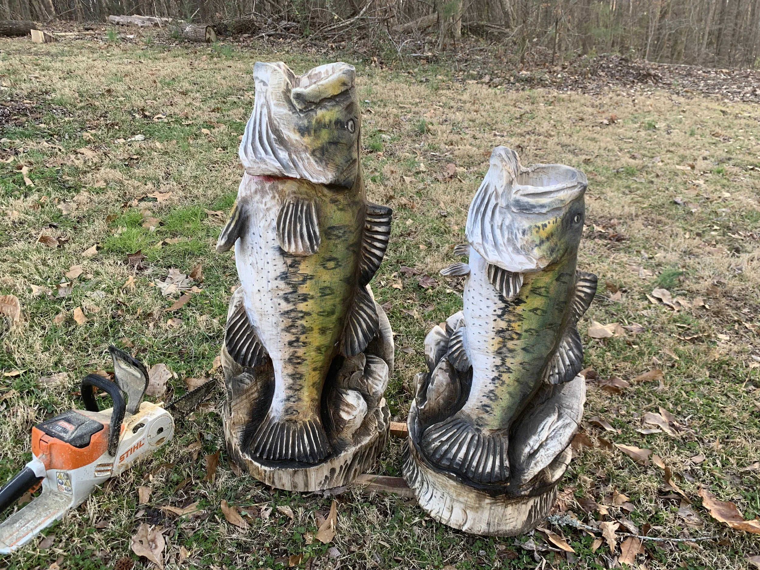 Carved wooden fish sculptures arranged outdoors on grass and surrounded by fallen leaves, with a chainsaw lying on the ground nearby.