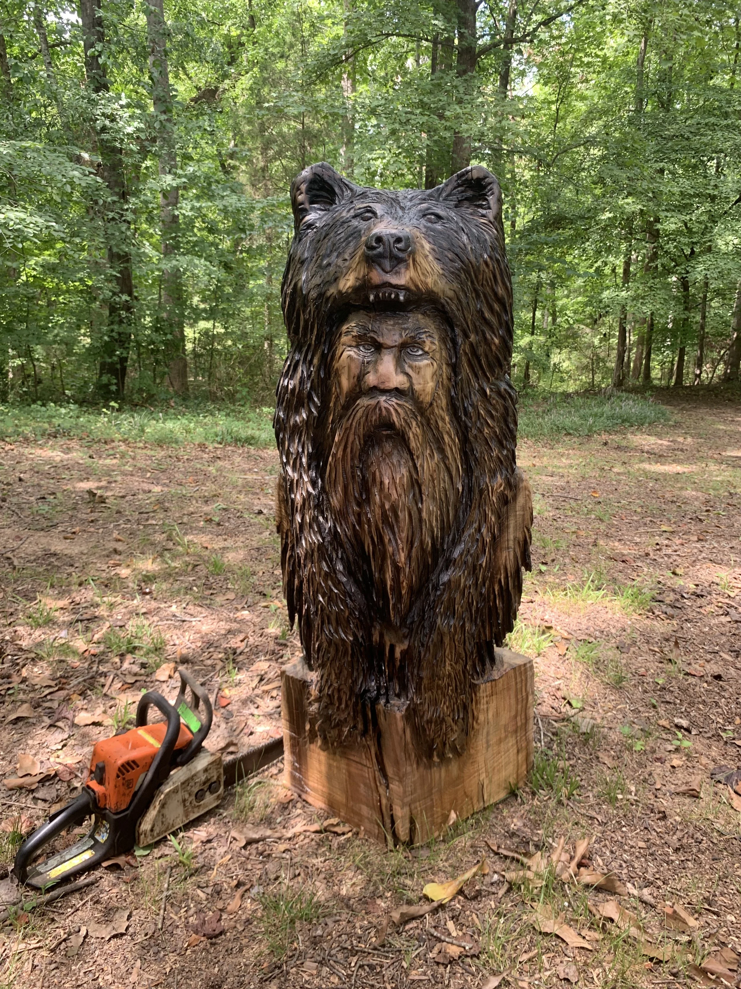 A wooden sculpture of a bear's head and an intense-looking human face, carved from a tree trunk, outdoors in a green wooded area. A chainsaw is on the ground nearby.