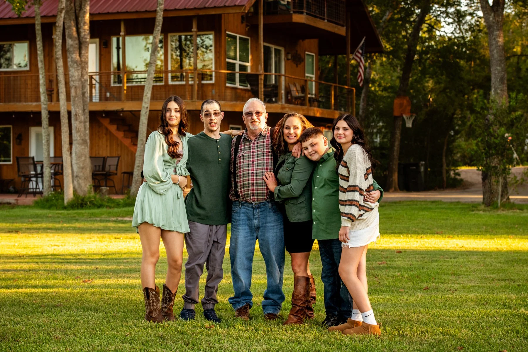 Family of six standing outdoors in front of a wooden house, smiling and posing for the photo.