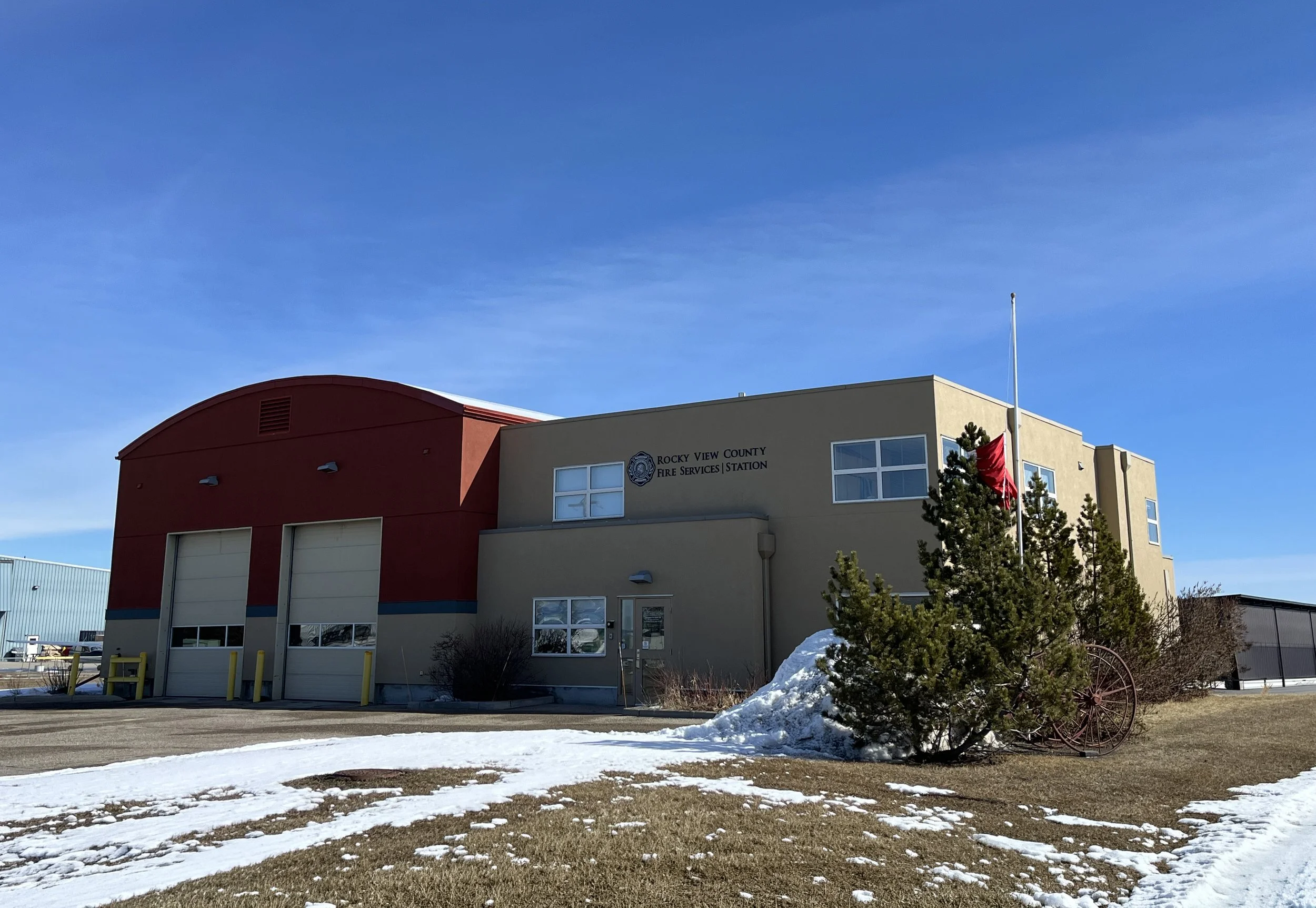 Image of a fire station building with a red curved roof and a beige section, with the sign 'Rocky View County Fire Services Station' and a Canadian flag flagpole nearby, under a clear blue sky with patches of snow on the ground.