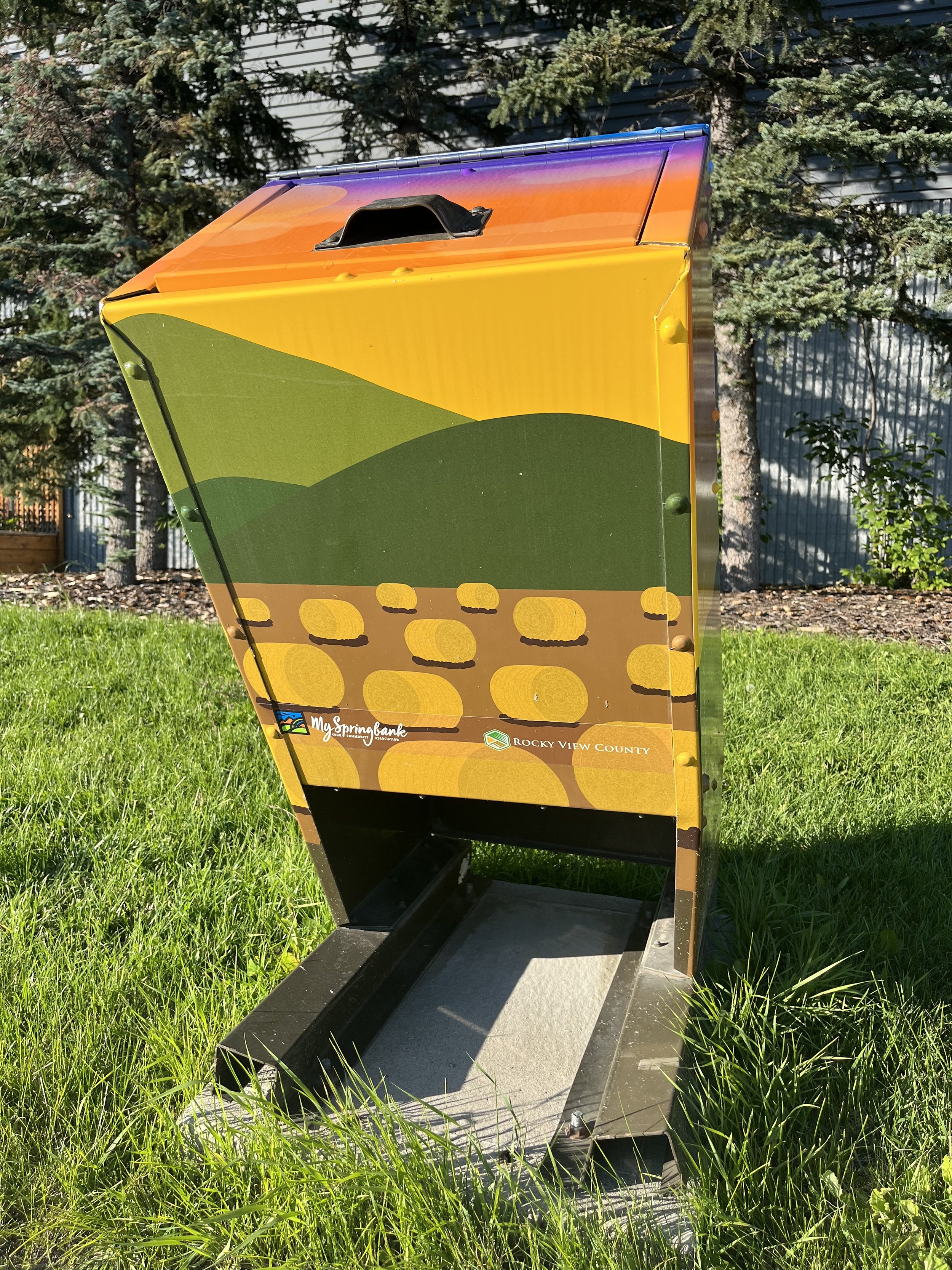 Colorful mailbox painted with a landscape scene featuring green hills, yellow and orange sky, and brown ground with yellow rocks, in an outdoor setting with grass and trees.