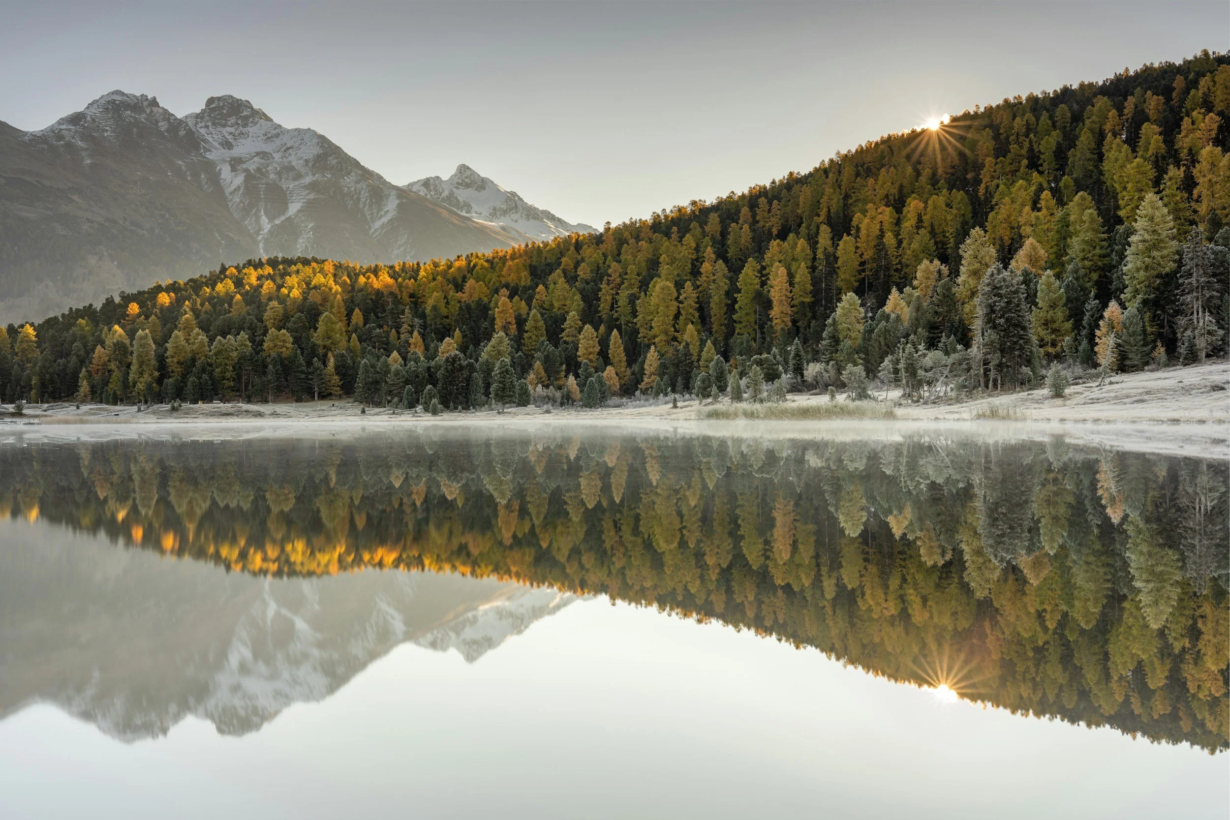 A scenic mountain landscape with snow-capped peaks, a forest of trees showing fall colors, and a calm lake reflecting the scenery, with the sun shining over the treetops.