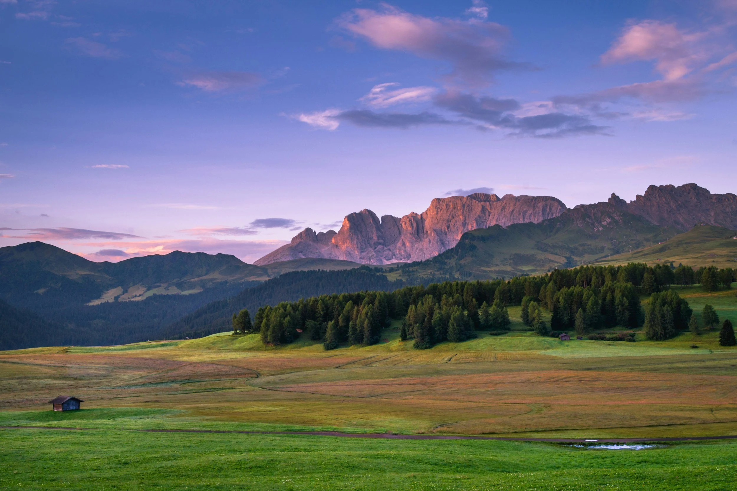 Scenic landscape of mountains at sunset with rolling green fields and a small wooden barn in the foreground.