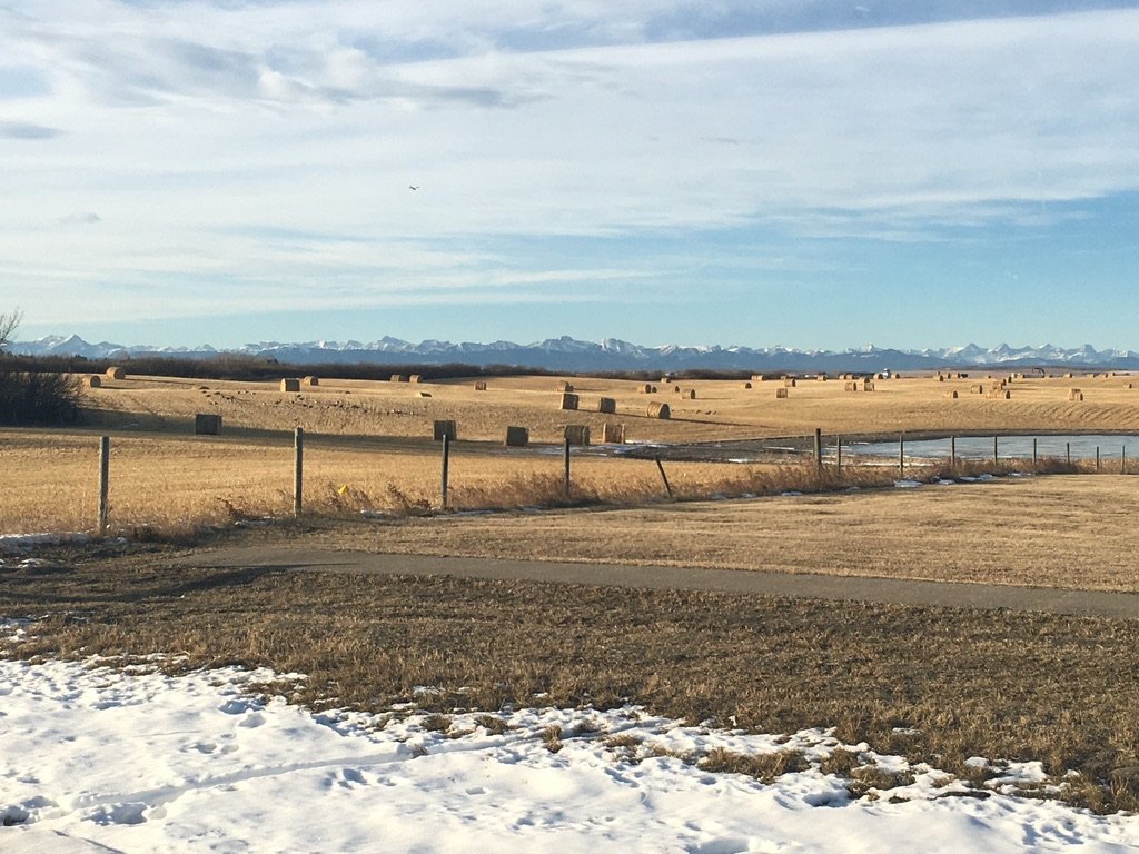 Open field with hay bales, snow patches, wooden fence, body of water, and distant snow-capped mountains under a partly cloudy sky.