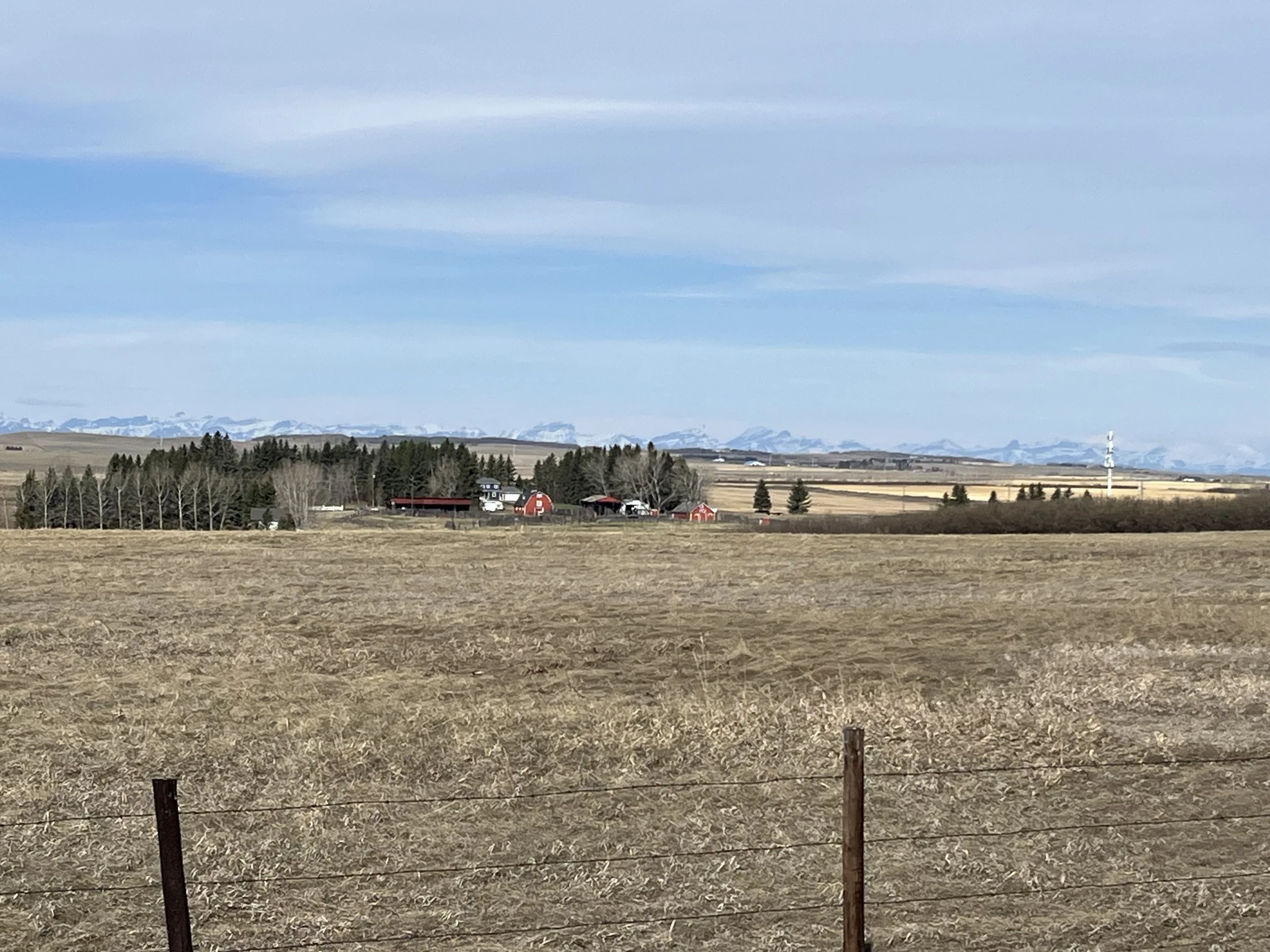 Wide rural landscape with dry fields in foreground, farm buildings, trees, and distant mountains under a blue sky with clouds.