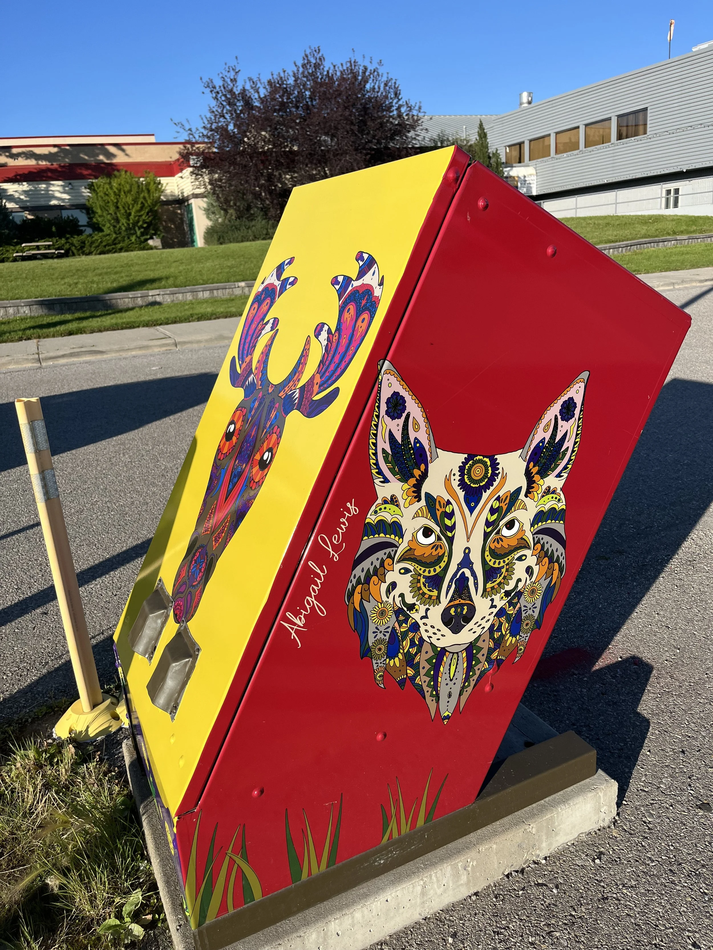Colorful public art box featuring a stylized deer and a stylized dog with intricate patterns, set outside on a sidewalk with a grassy area and modern buildings in the background.