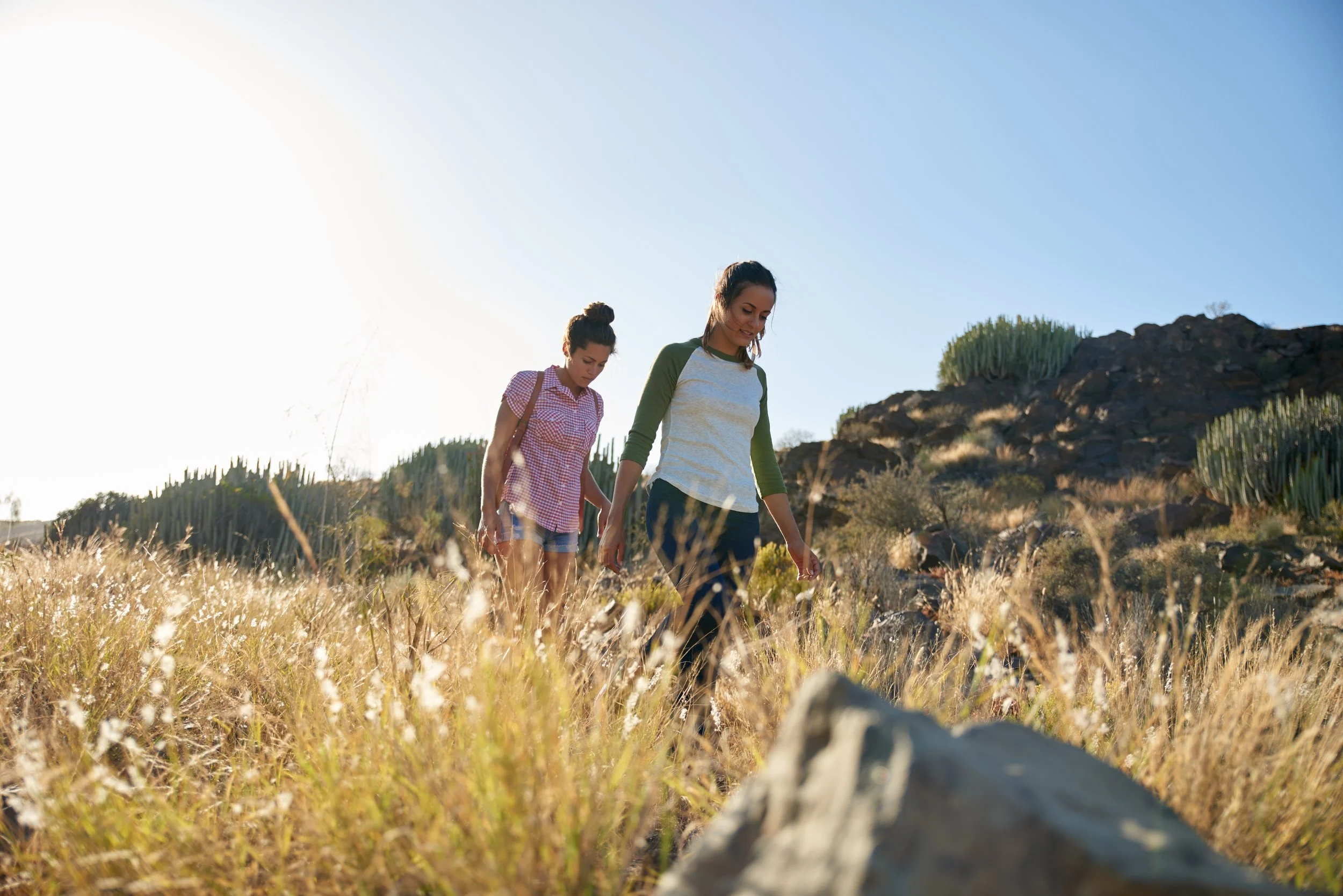 Two women walking through a dry grassy hillside with cacti and rocks, under a clear blue sky.