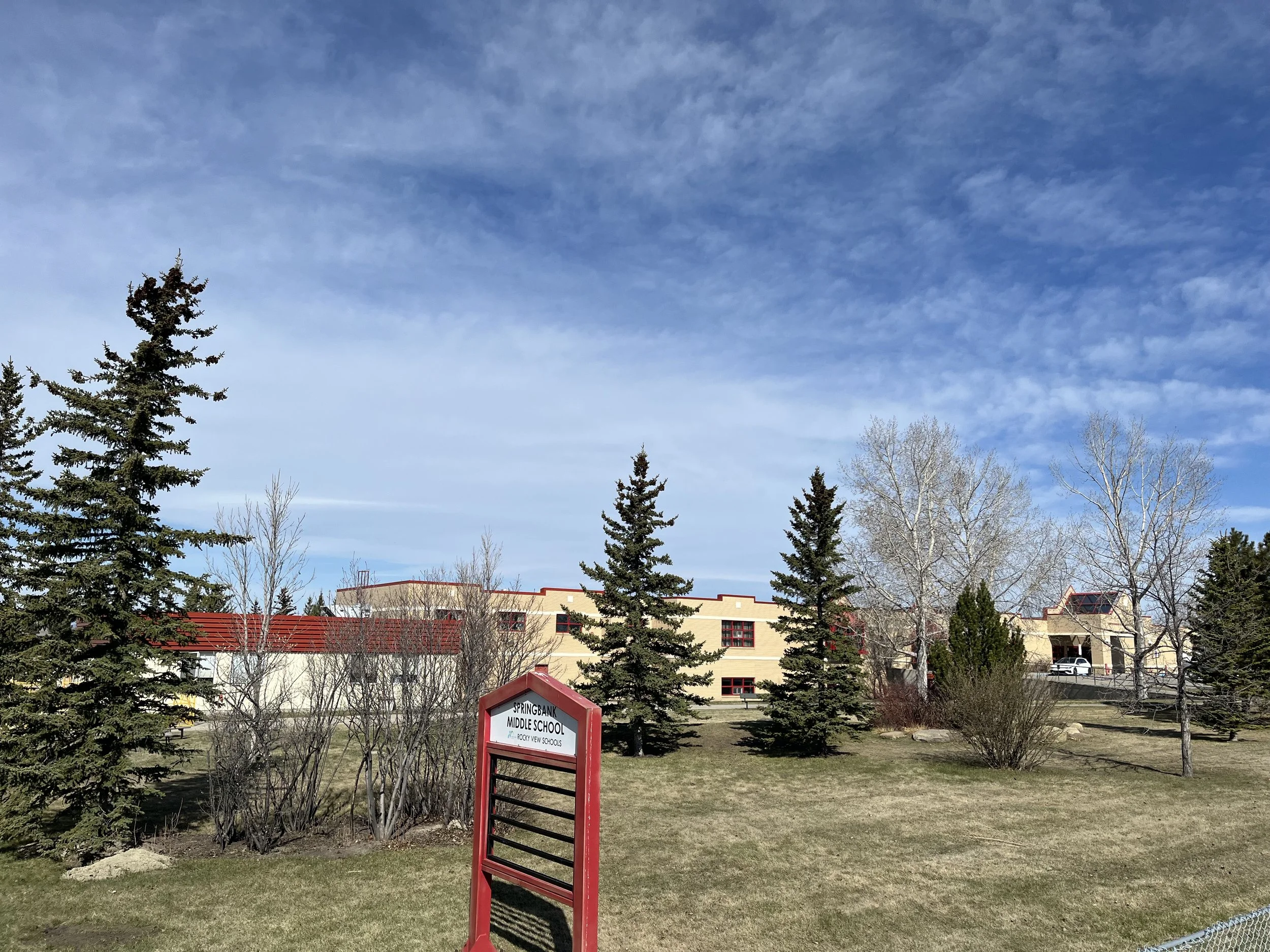 View of Springbank Middle School with a sign in the foreground, several trees, and a building with red-roofed sections under a cloudy sky.