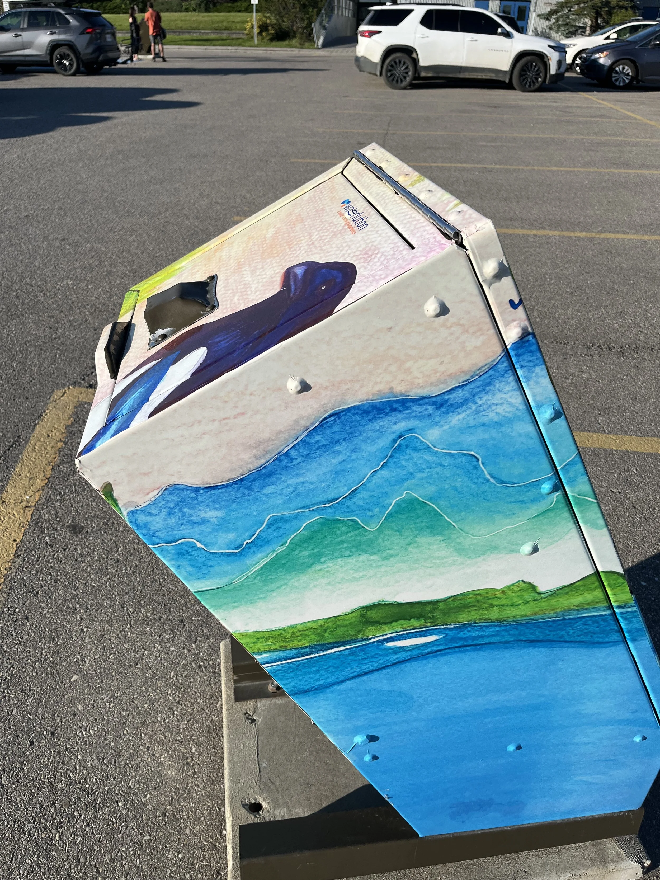 A colorful painted landscape scene on a large rectangular object, possibly a piece of artwork or outdoor decor, positioned in a parking lot with cars and trees in the background.