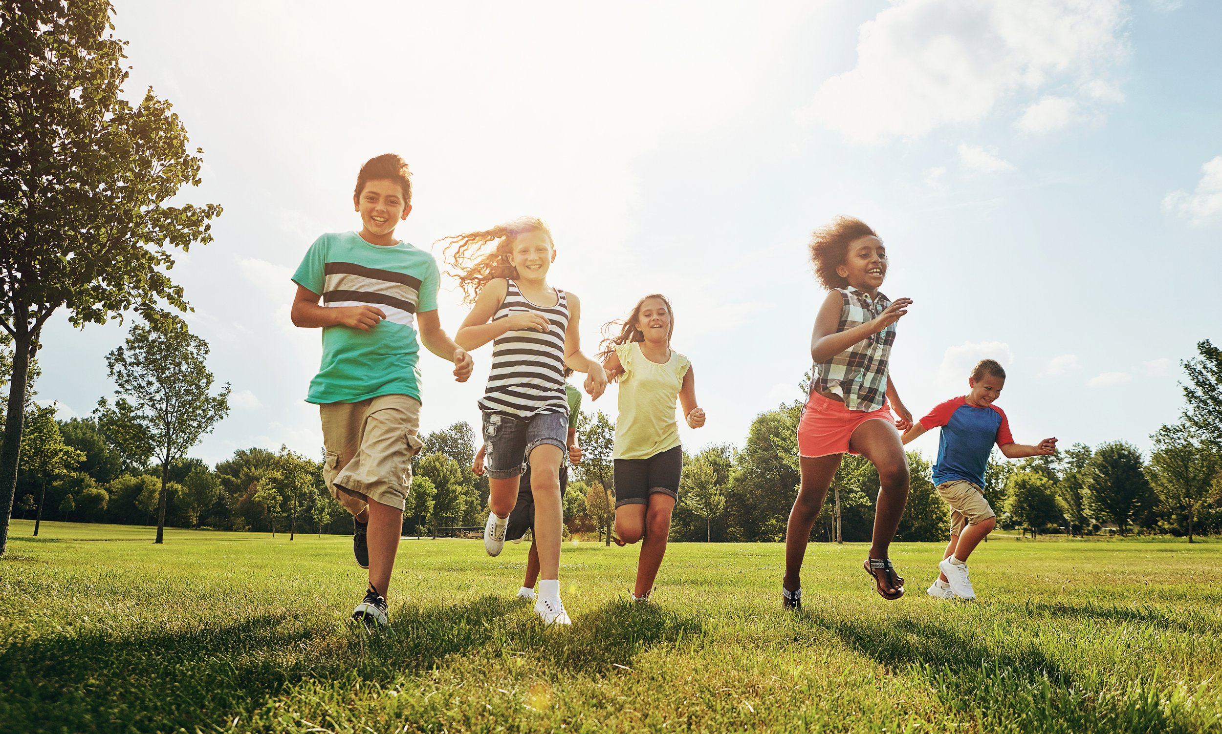 Children running and playing in a park on a sunny day with green grass and trees in the background.