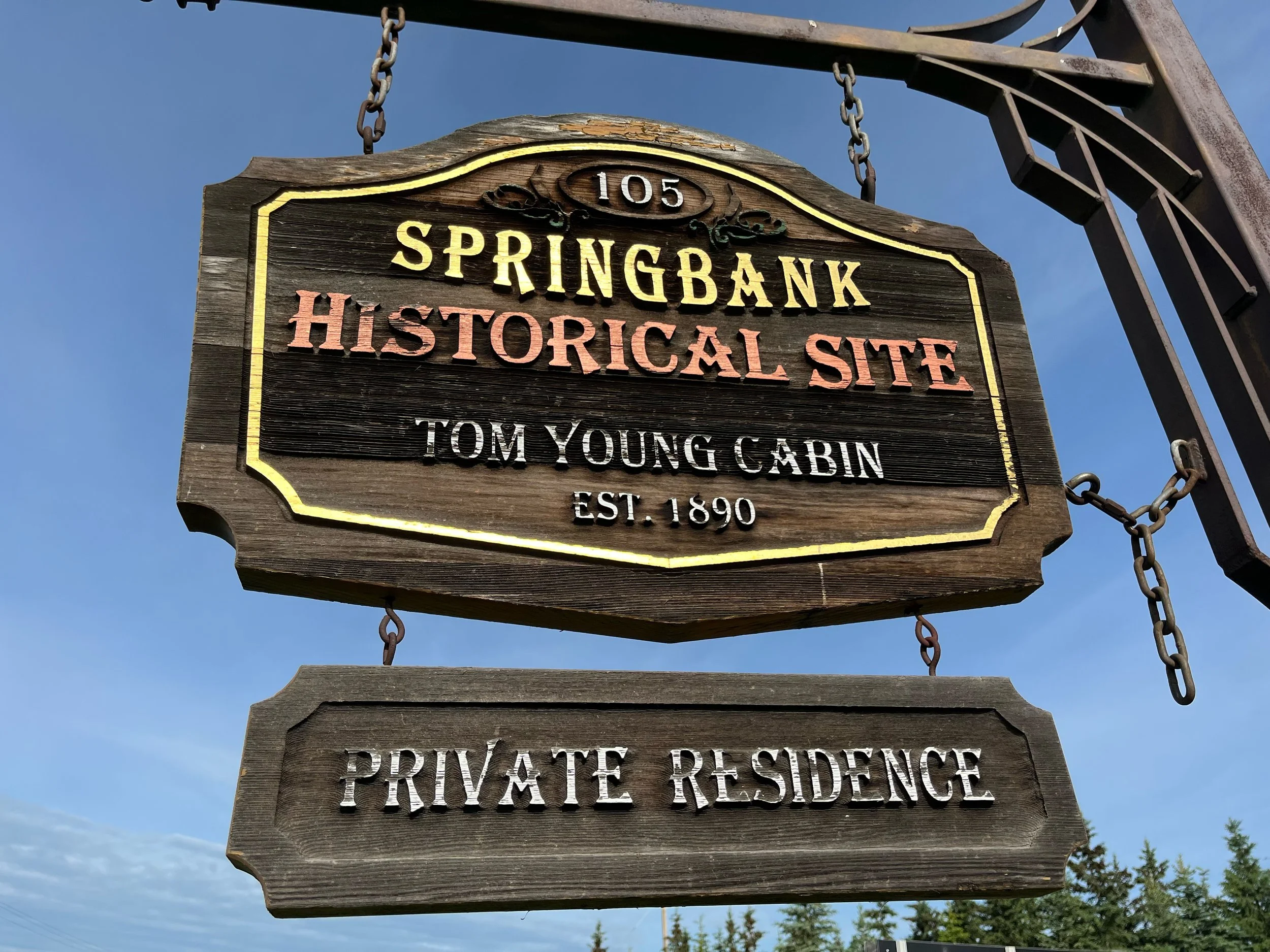 Wooden sign for Springbank, a historic site, with chains attaching it to a metal frame against a blue sky with some clouds.