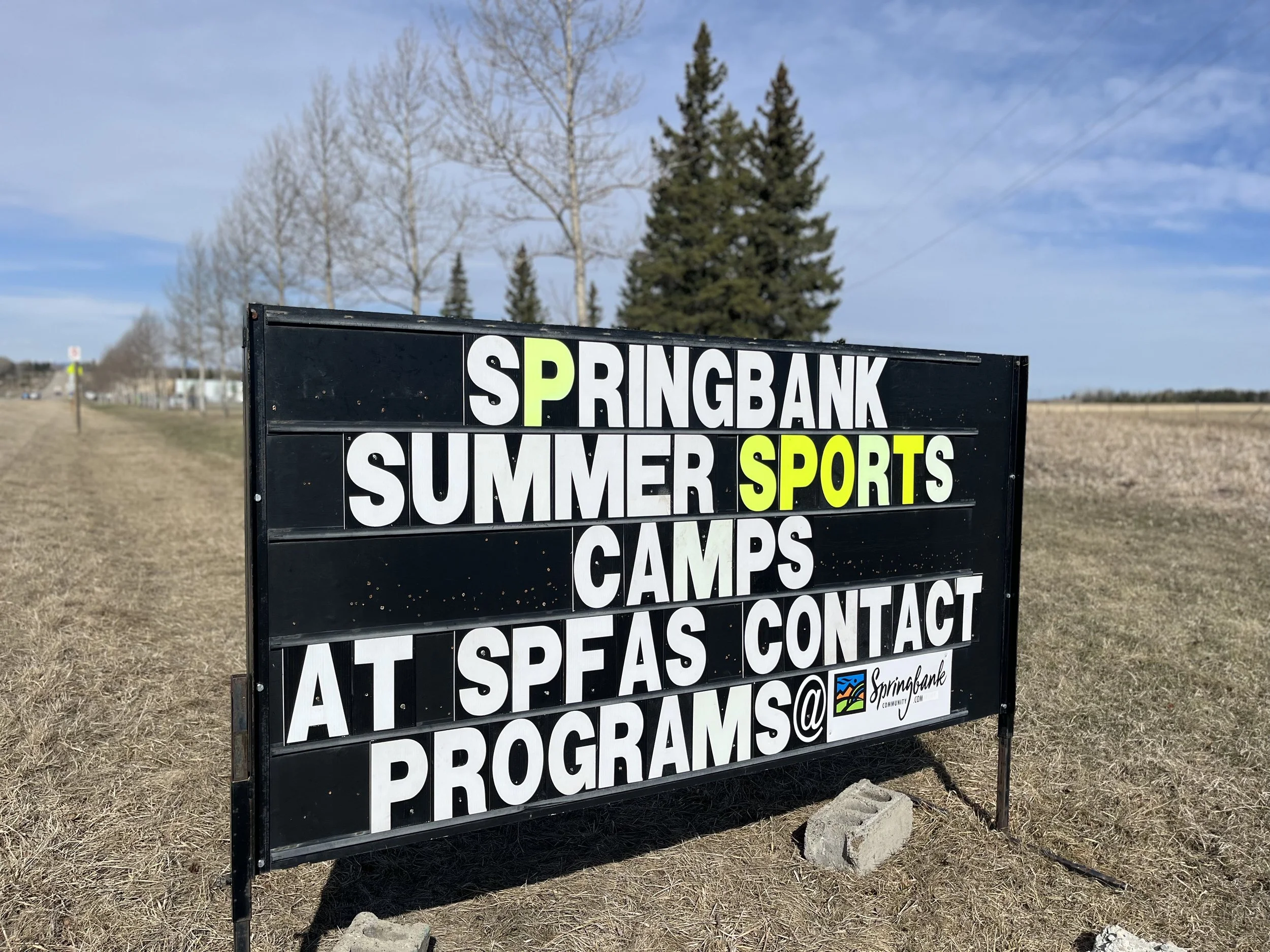 A black signboard with white and yellow text outdoors on a cloudy day, advertising summer sports camps at Springbank Community.