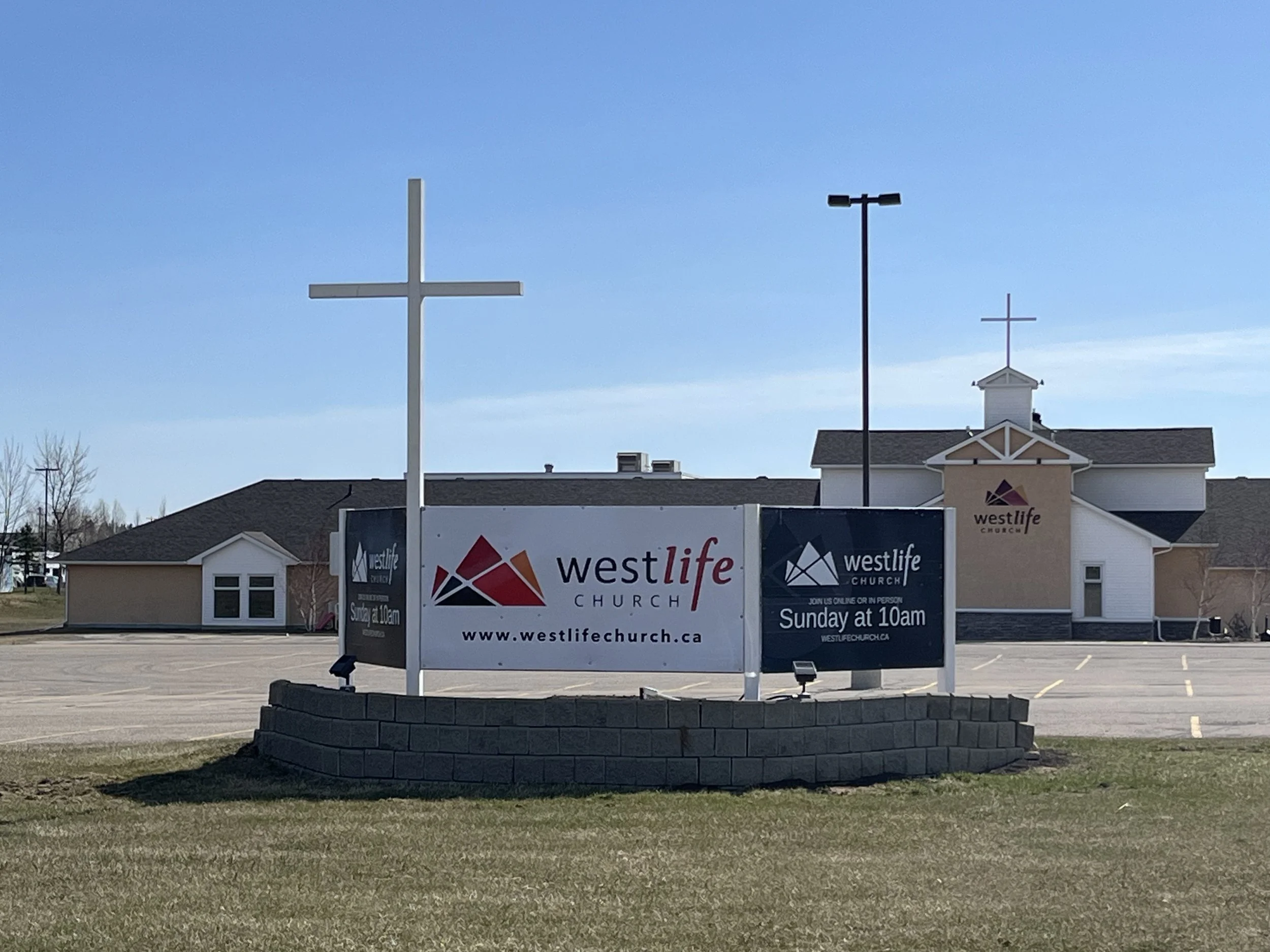 Church building with cross on roof, advertising signs for Westlife Church in parking lot, clear blue sky.
