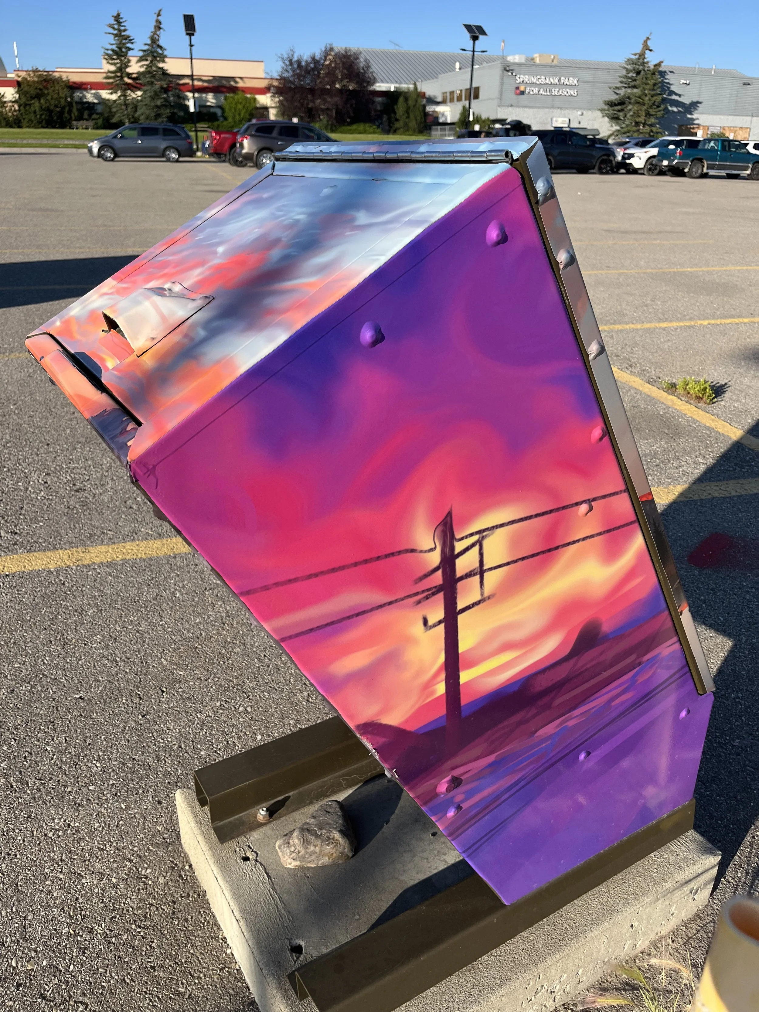 A colorful painted utility box in a parking lot with a sunset or sunrise sky and power lines painted on it.
