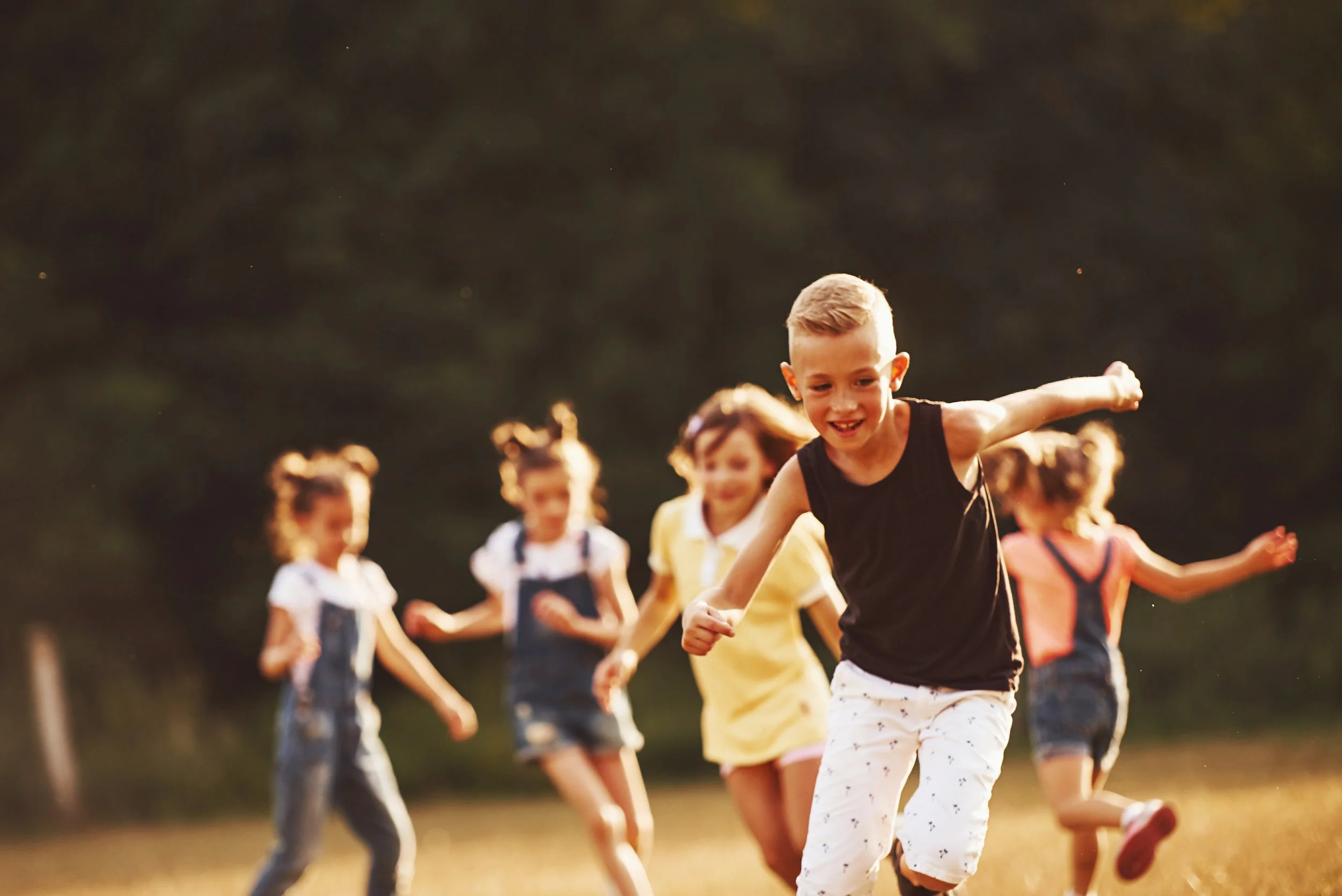 A group of children running and playing outdoors in a park or field during daylight, with trees in the background.