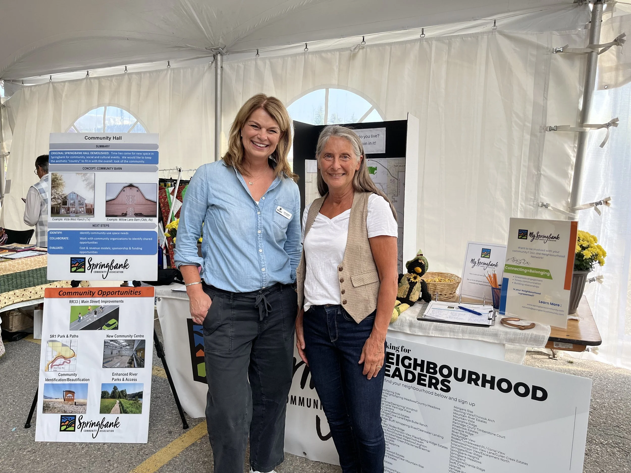 Two women smiling at an outdoor community event, standing in front of informational display boards about neighborhood improvement projects.