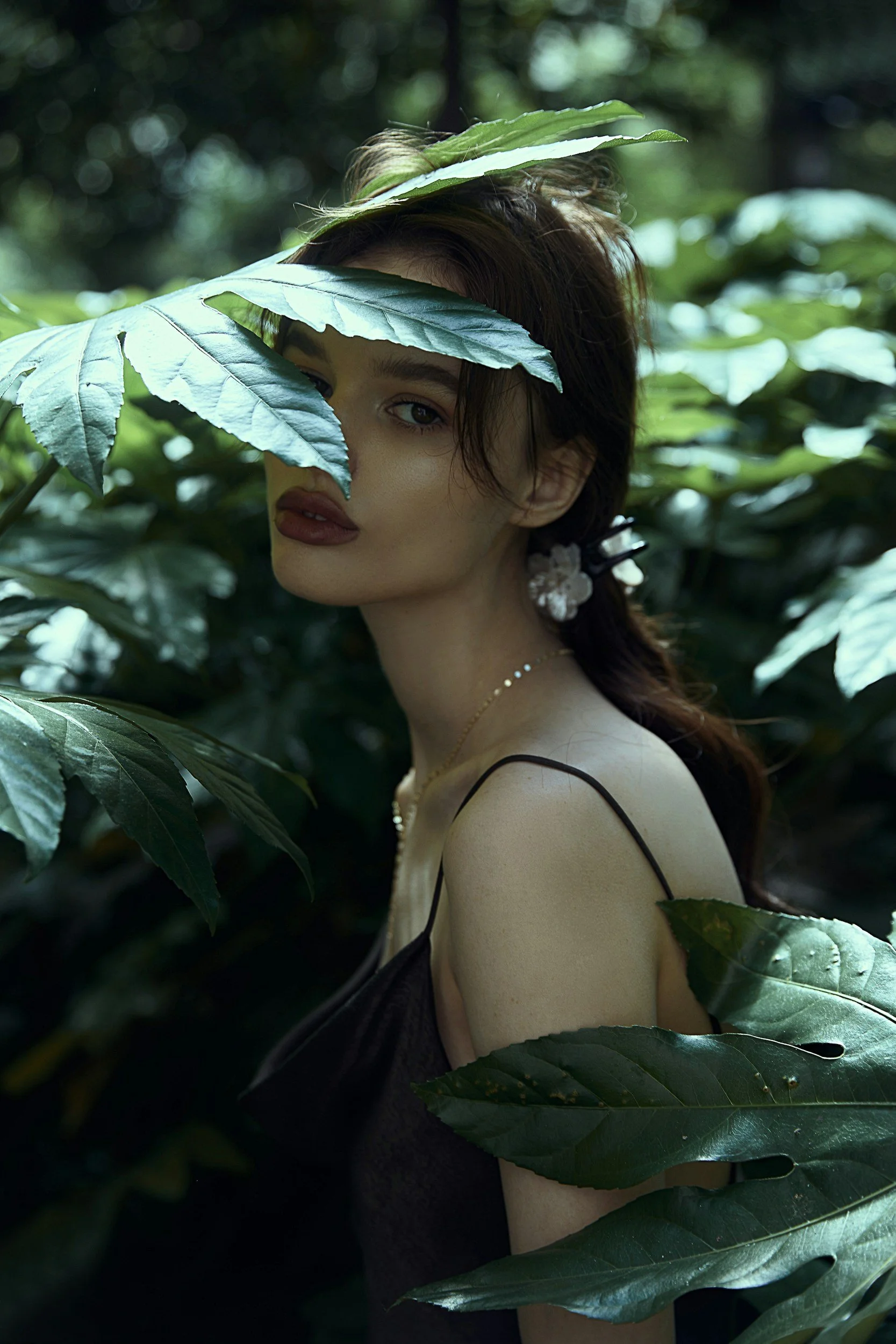 A woman with makeup and earrings poses amidst large green leaves, partly covering her face, in a lush, natural setting.