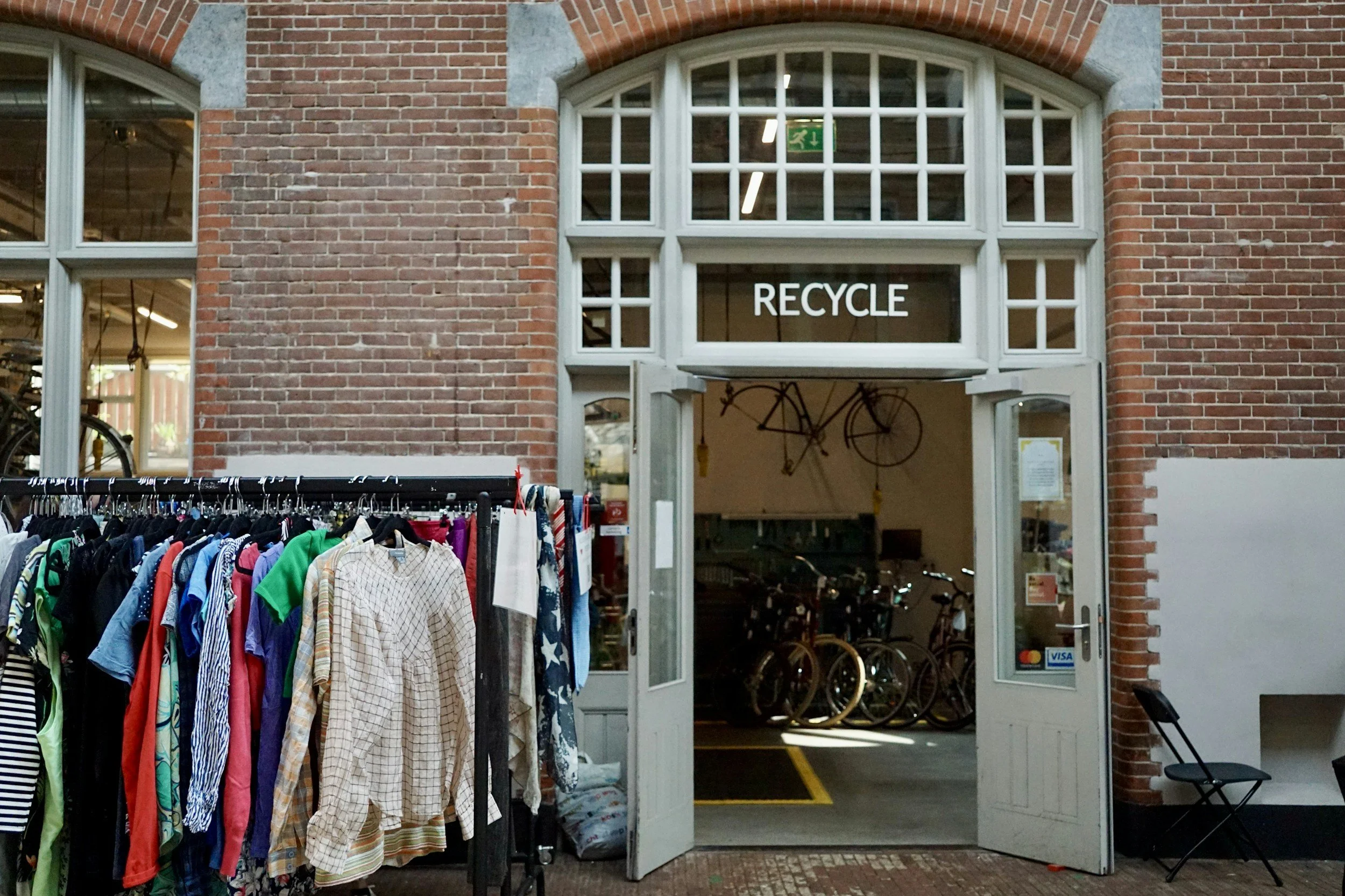 Entrance of a thrift store with a clothing rack outside, bicycles hanging inside, and a sign that says recycle.