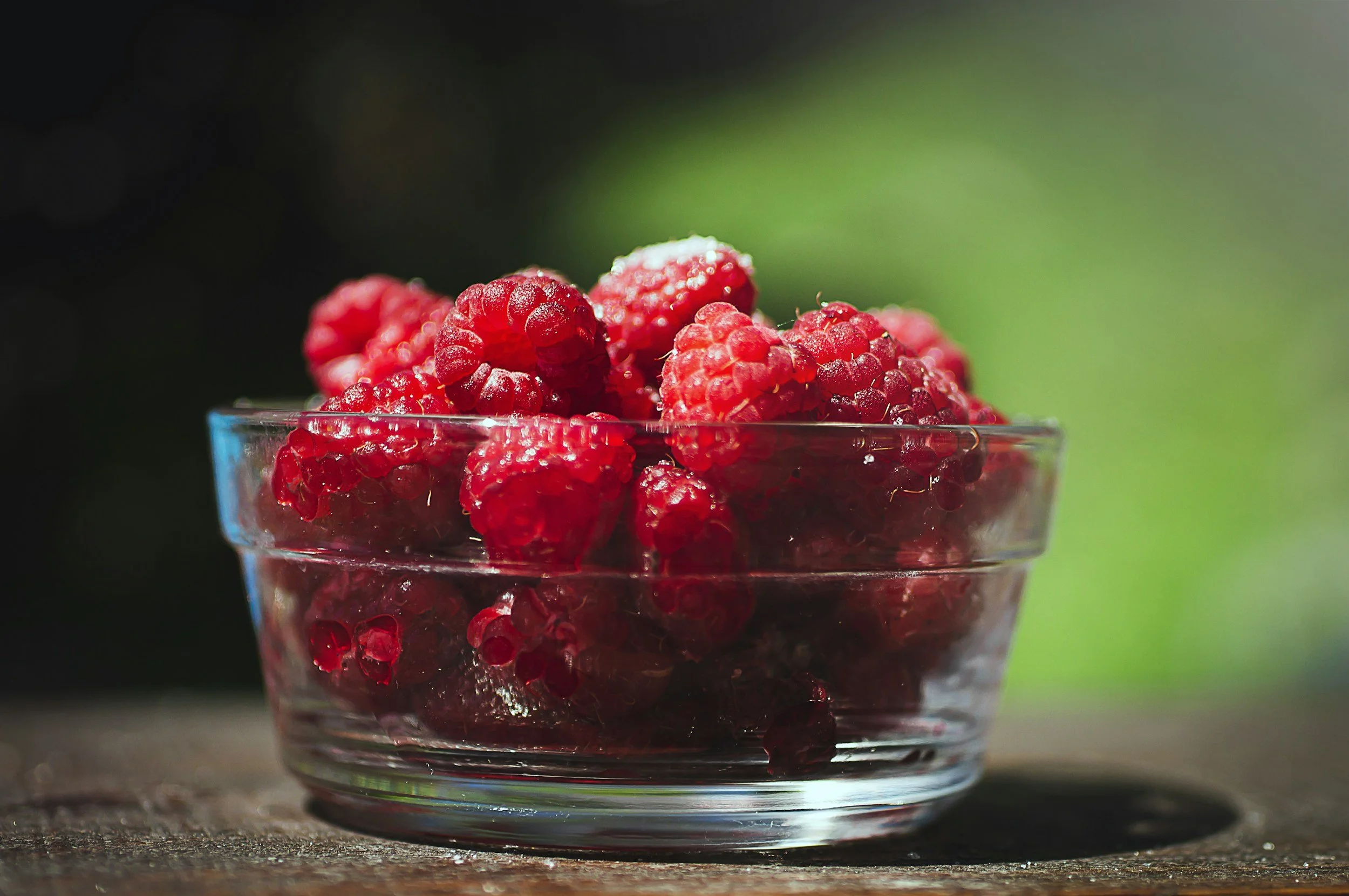 A glass bowl filled with fresh red raspberries on a wooden surface with a blurred green natural background.