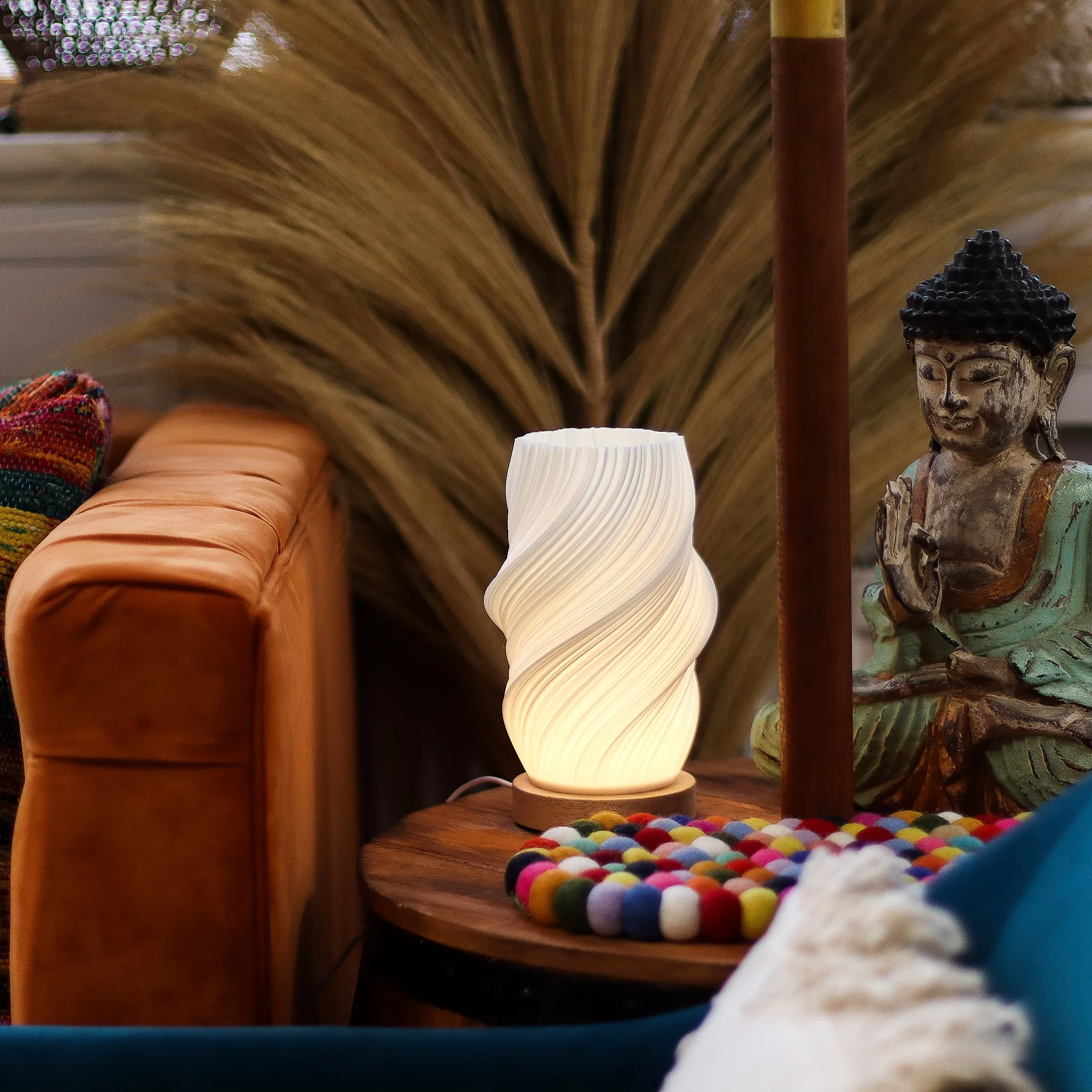 A cozy living room corner with a glowing white lamp on a round wooden side table, next to a colorful beaded mat, a sculpture of a seated figure with a headdress, and a multicolored woven cushion on a brown velvet sofa, with dried palm leaves in the background.