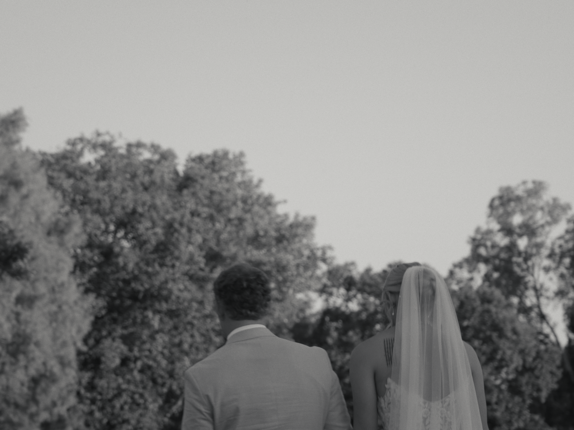 A black and white photograph of a man and woman dressed in formal attire, walking together outdoors near trees, the woman's veil covering her hair.
