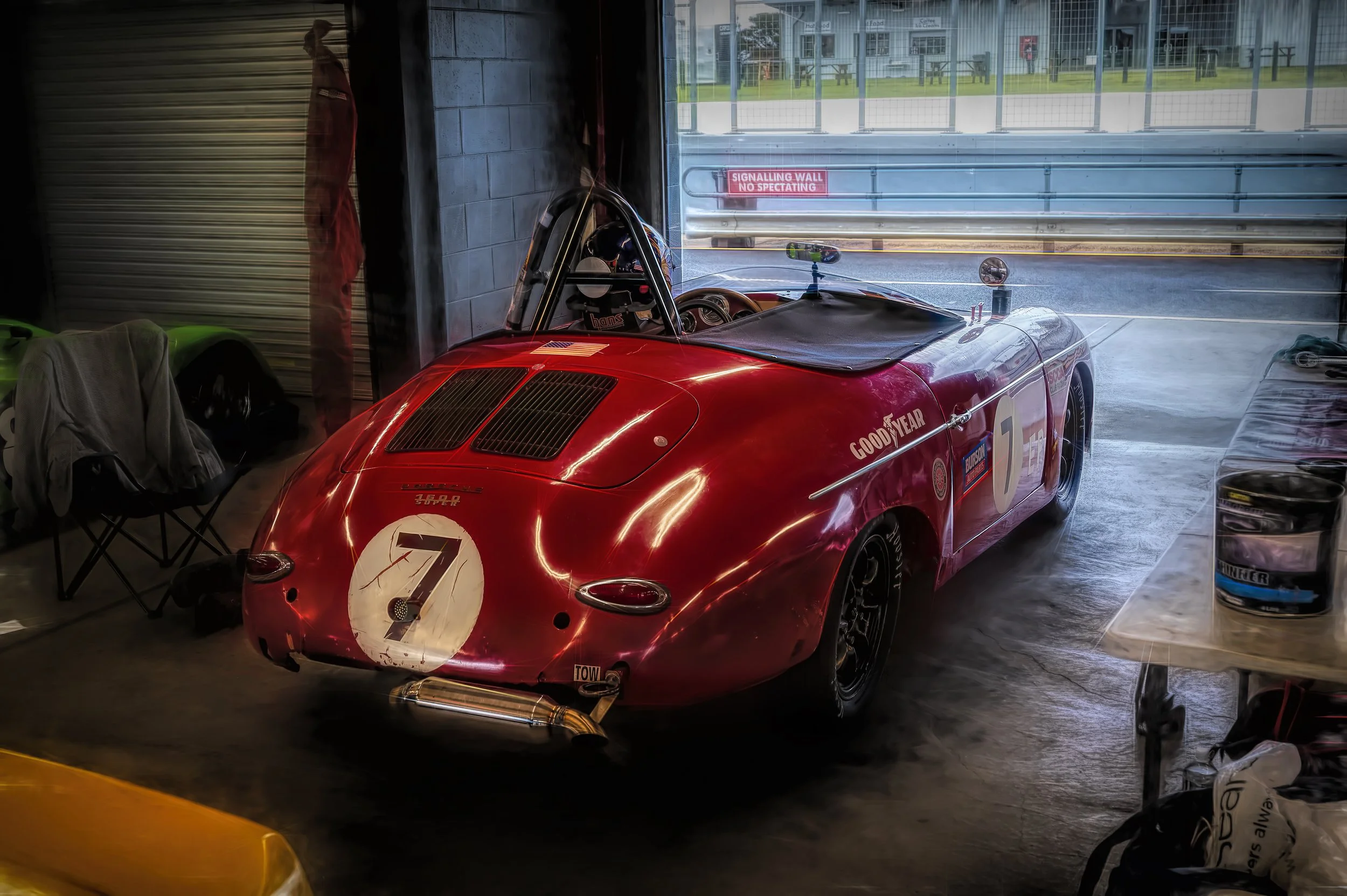 A vintage red Porsche speedster, parked inside a pit garage with a view of a racetrack through the open door.