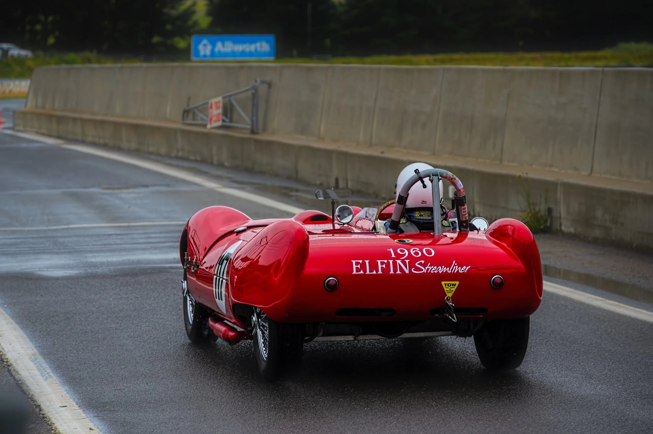 A red vintage race car driving on a wet road with a concrete barrier on the side and an overhead road sign in the background.