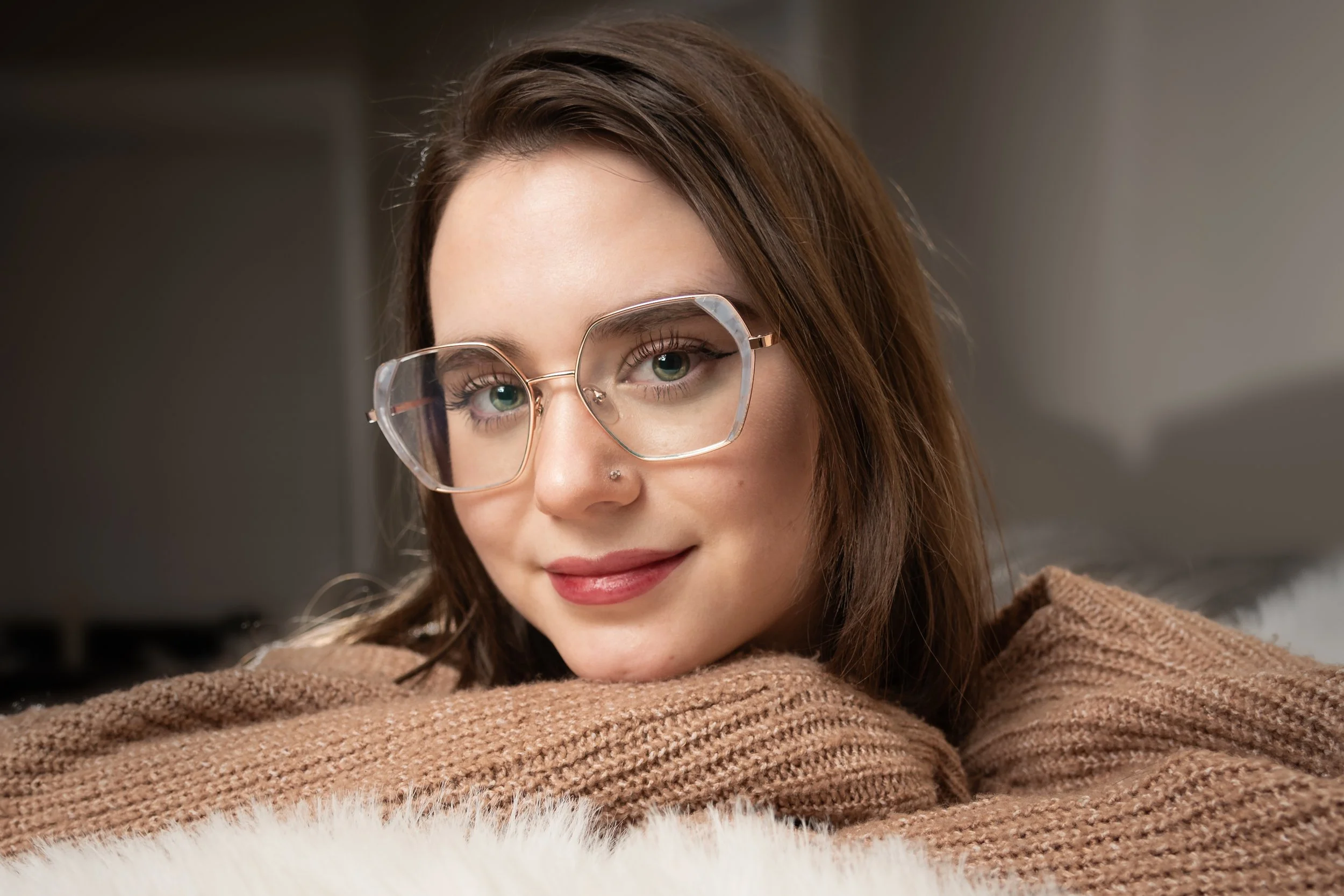 Close-up of a young woman with light skin, hazel eyes, brown hair, wearing clear-framed glasses, resting her chin on a soft brown blanket with a cozy indoor background.
