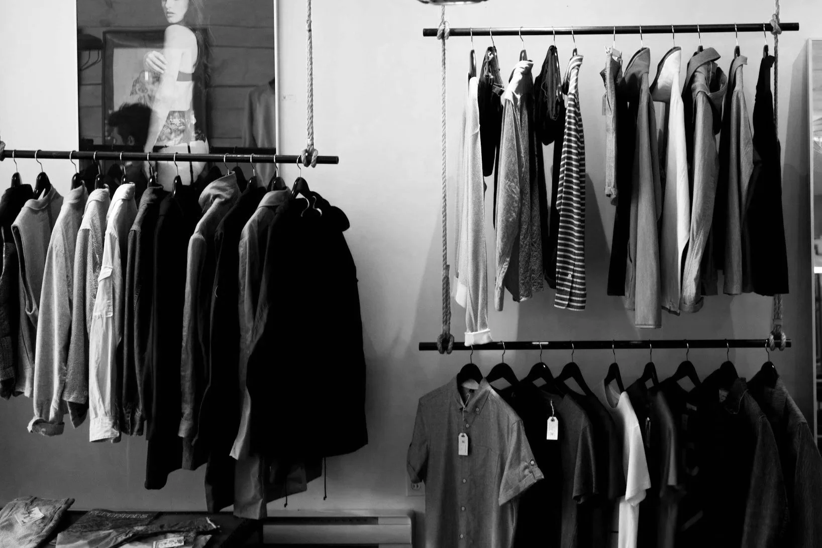 Clothing store display with shirts and jackets on racks, a mirror reflecting a woman, and a table with folded clothes.