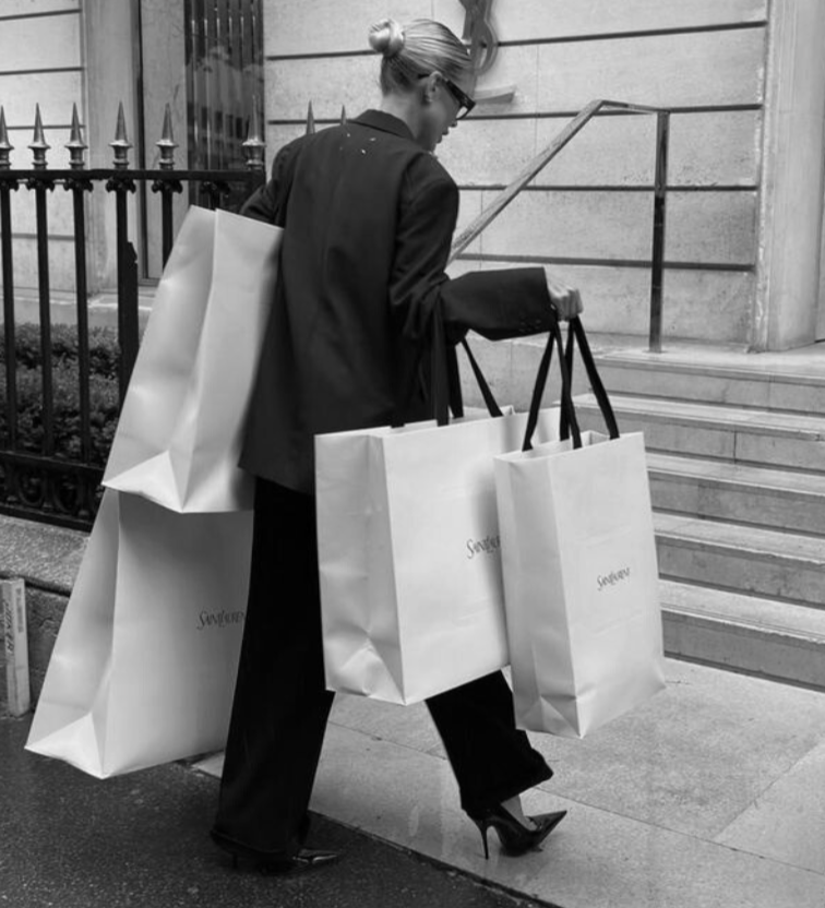 A woman in a blazer and high heels carries multiple shopping bags on a city sidewalk near stairs and a metal fence.